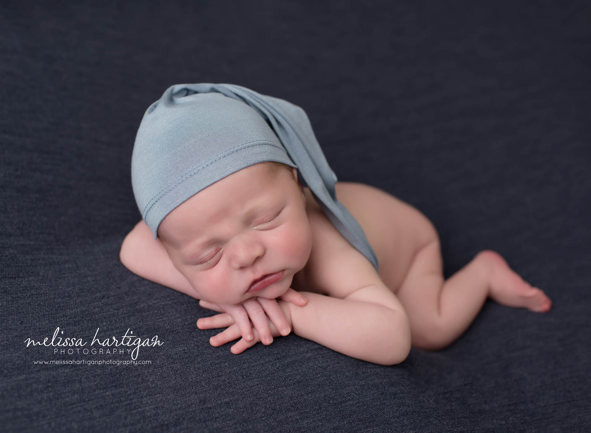 newborn baby boy posed on tummy with hands under chin wearing light blue sleepy cap