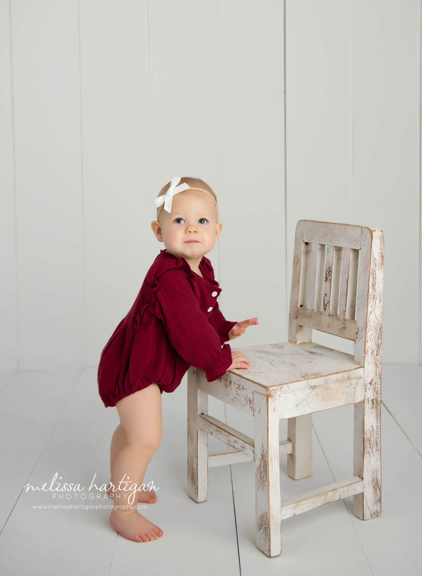 baby girl wearing red outfit and white bow in hair standing at chair milestone photography session