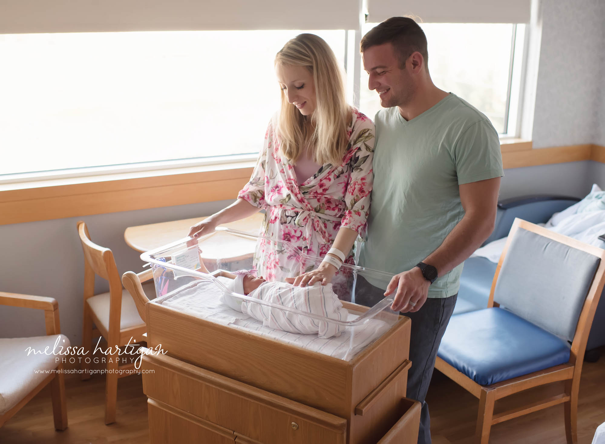 mom and dad standing over baby girl laying in hospital bassinet