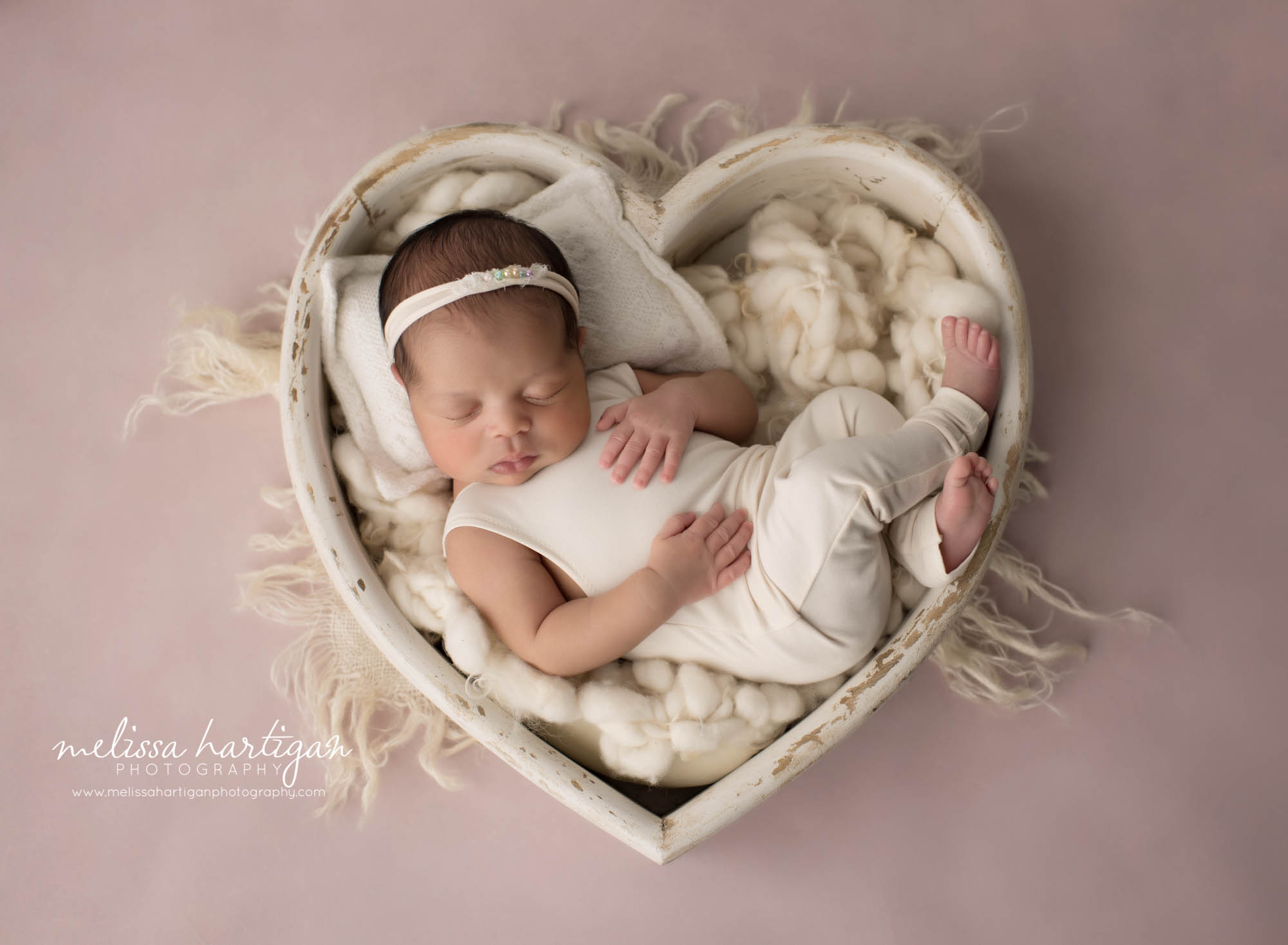 newborn baby girl posed in wooden heart bowl wearing cream baby girl outfit and headband