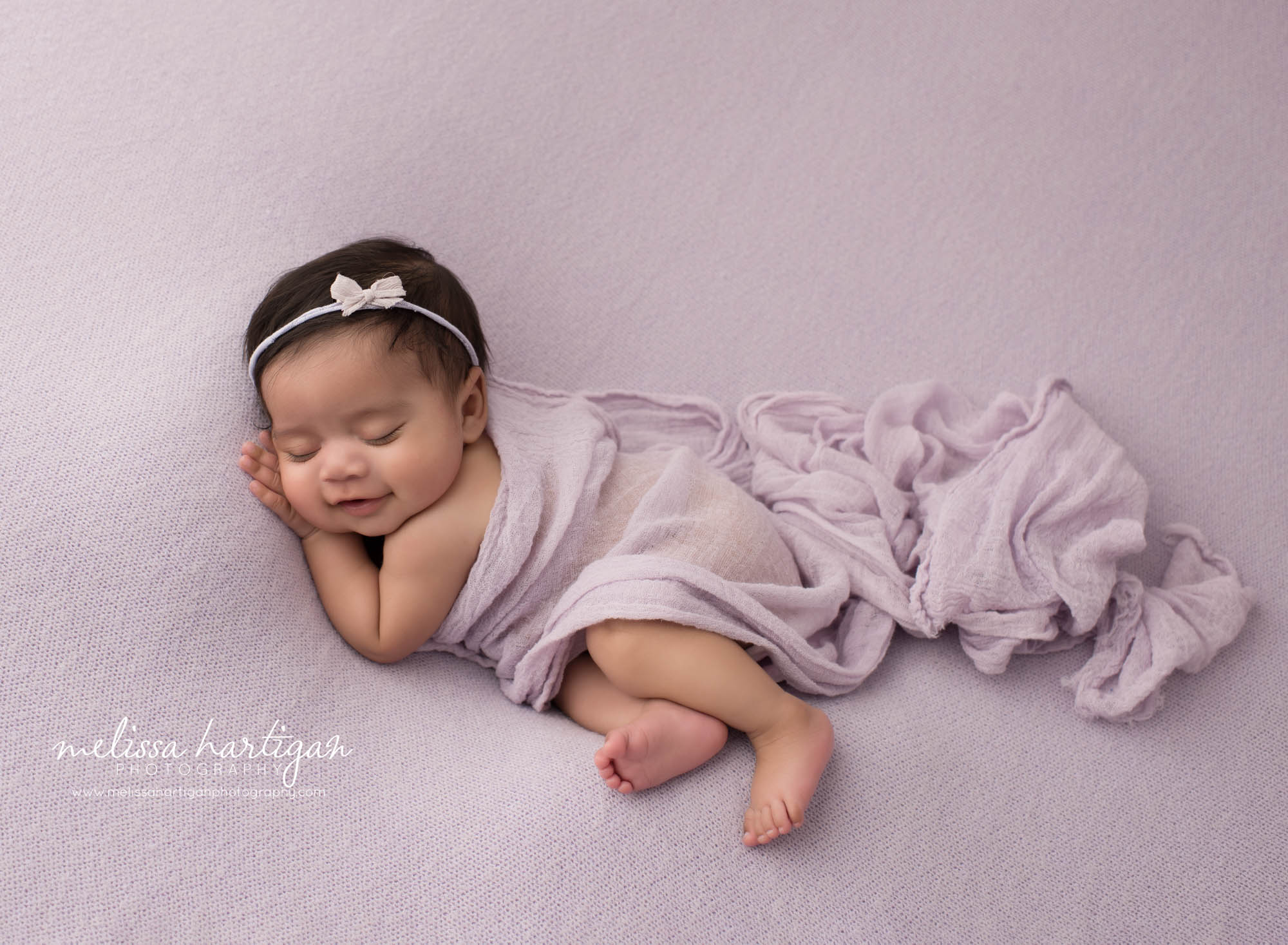 newborn baby girl posed on side sleeping smiling happy lavendar colored backdrop and wrap