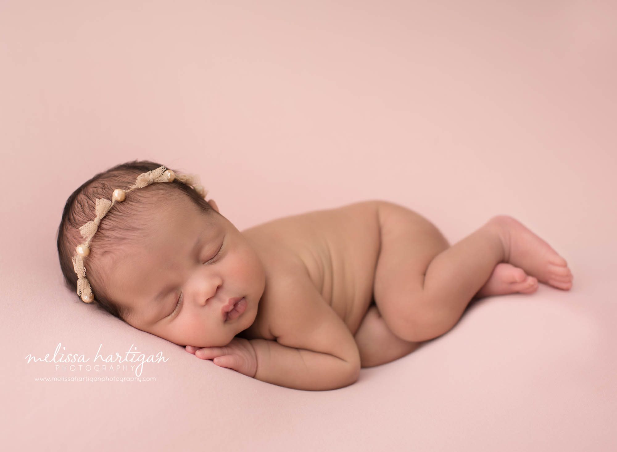 newborn baby girl posed on side on pink backdrop wearing gold beaded headband