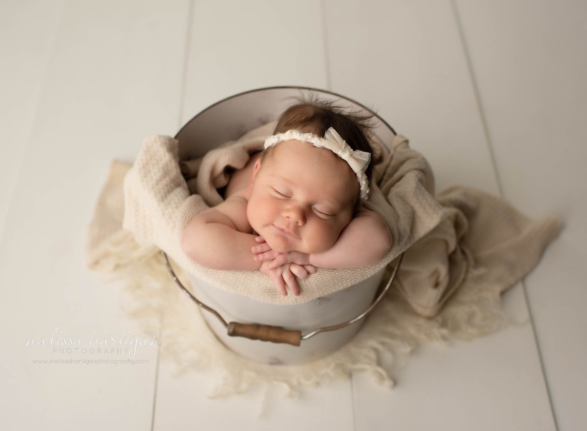 newborn baby girl posed in bucket with cream layer wrap and cream bow headband