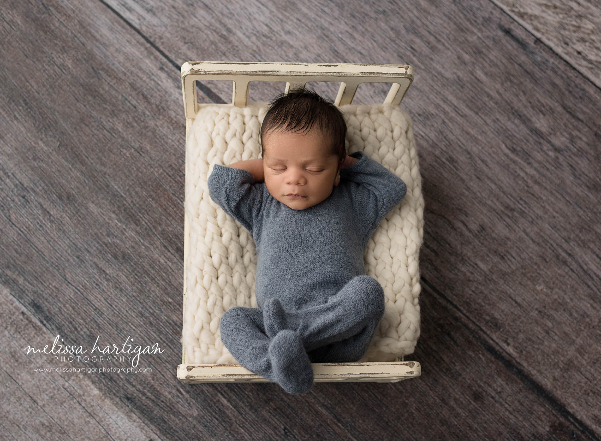 newborn baby boy posed on back sleeping with hands behind head on cream wooden bed prop and cream knitted chunky layer