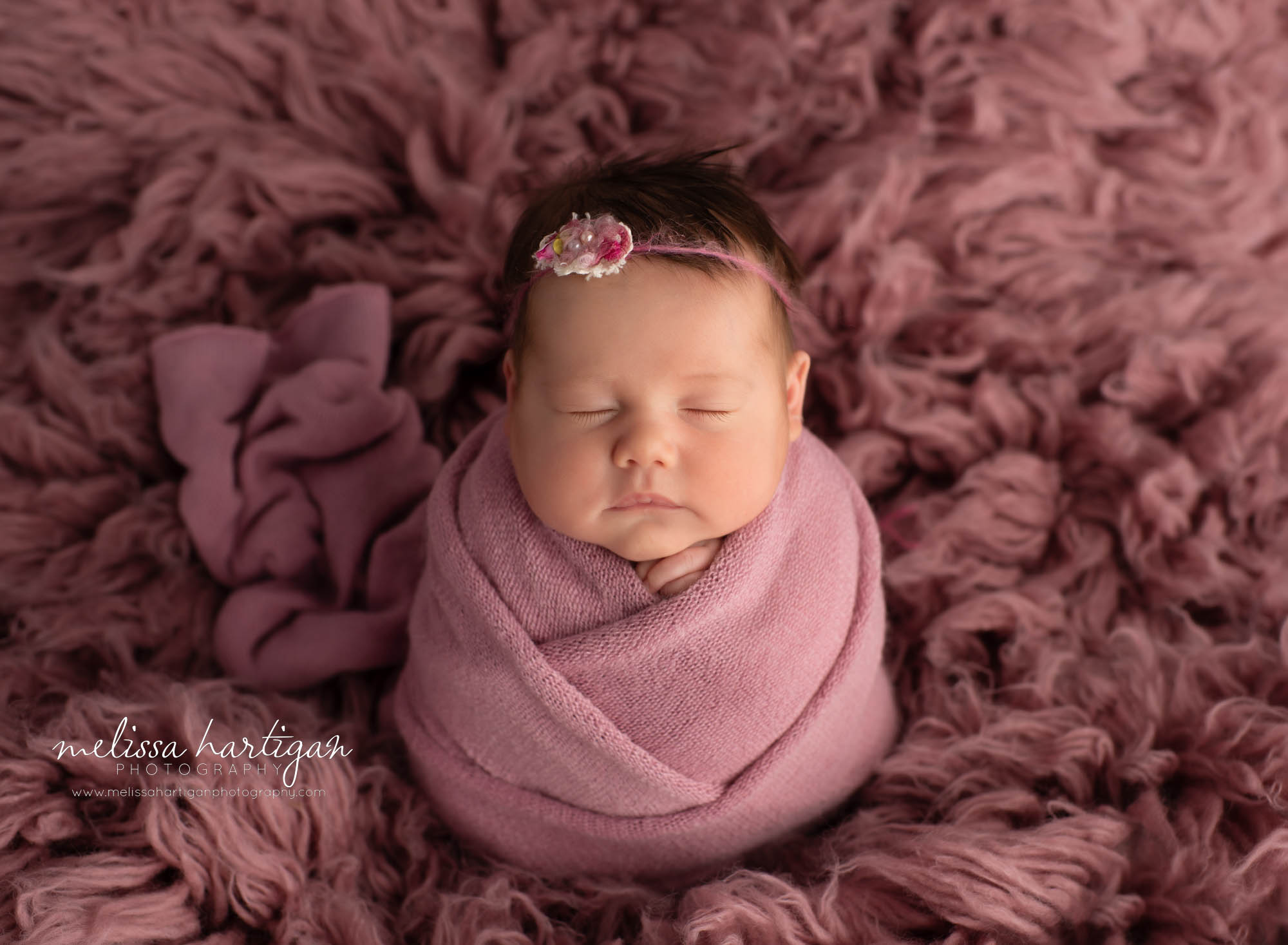 newborn baby girl swaddled in pink knitted wrap wearing headband tieback posed on dusty rose pink colored flokati