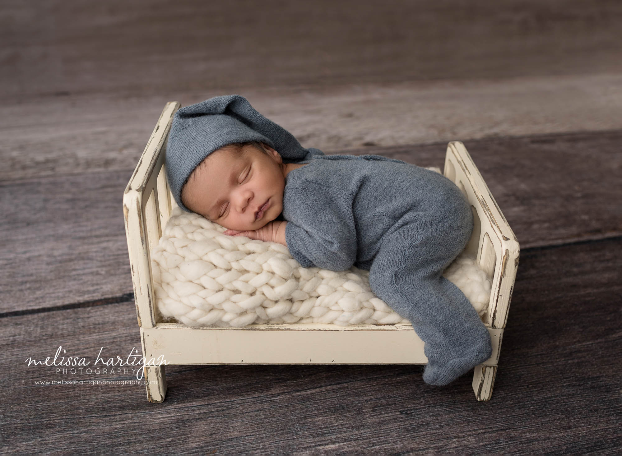 newborn baby boy posed on cream wooden bed prop wearing blue outfit and matching sleepy cap
