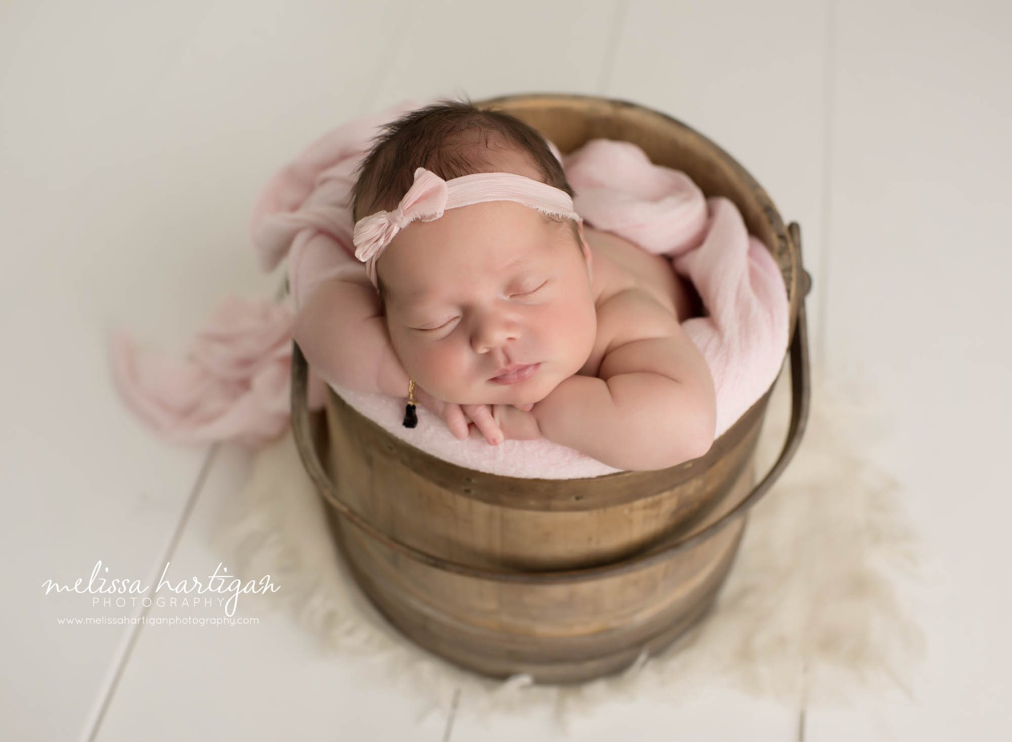 newborn baby girl posed in wooden bucket wearing pink bow headband and pink layer wrap