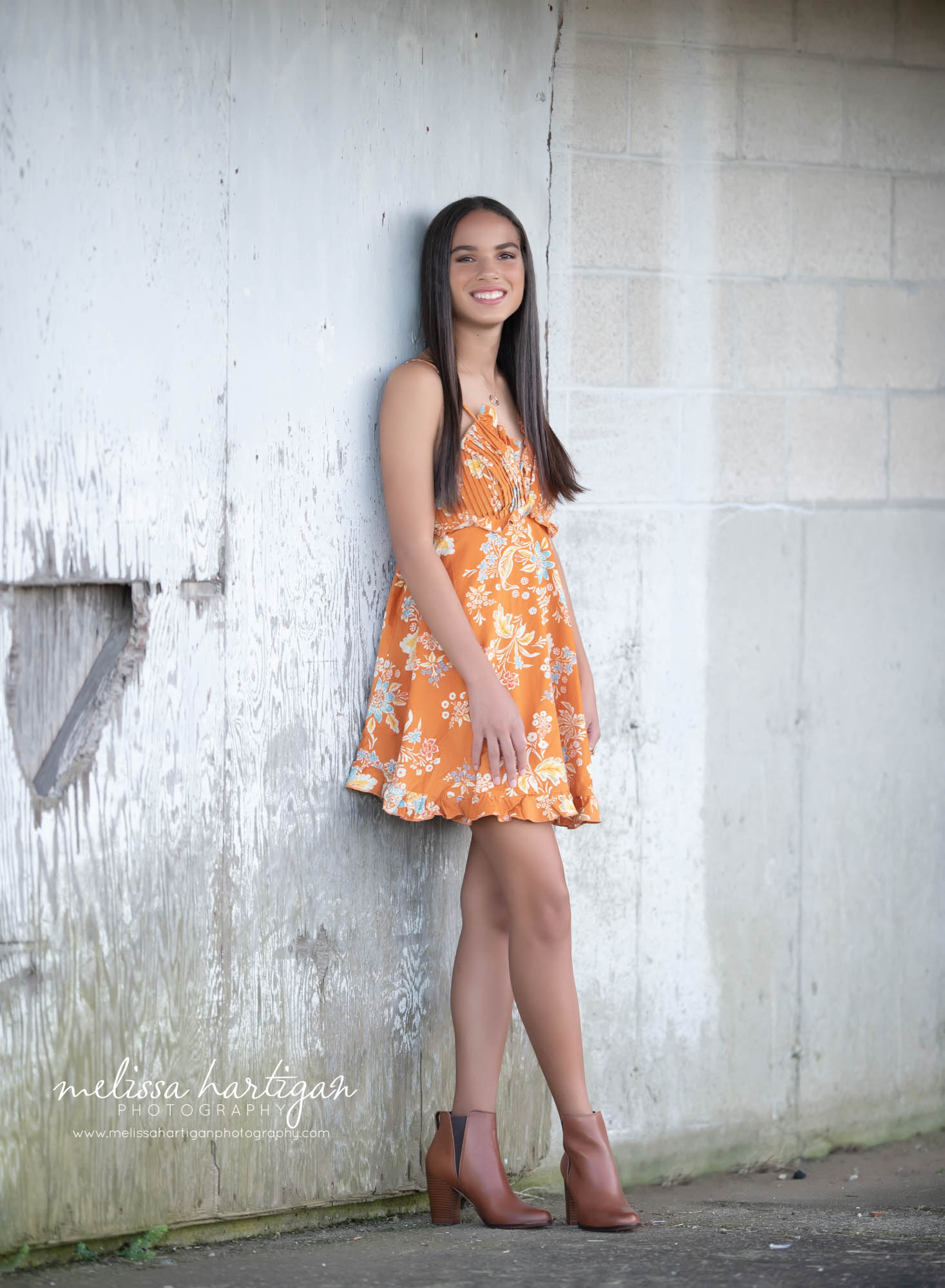girl wearing orange and yellow dress standing against wall cross legged celebrating high school graduation