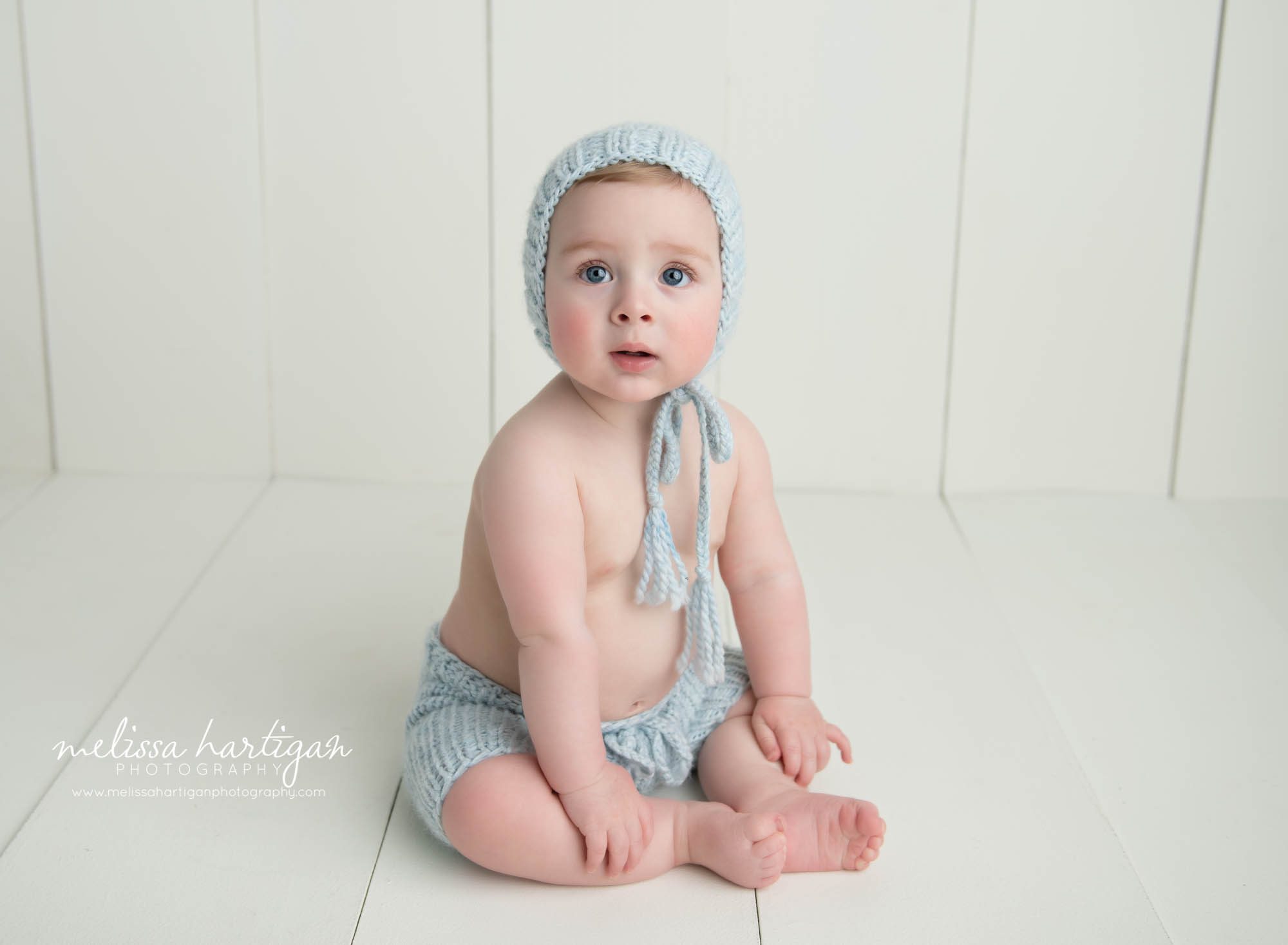 baby boy sitting on wooden boards in photography studio wearing light blue knitted bonnet