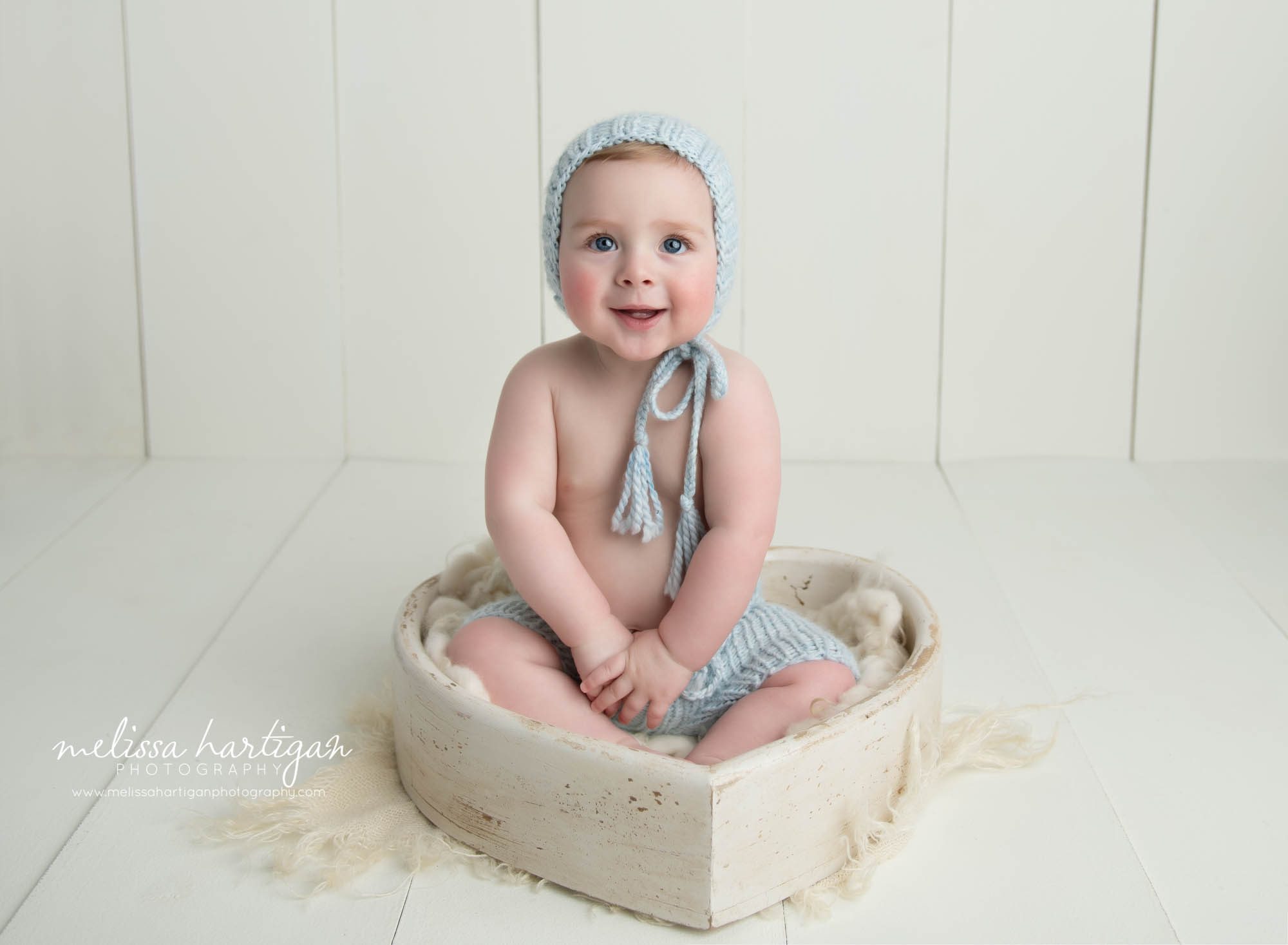 baby boy sitting in wooden heart bowl prop wearing blue knitted bonnet