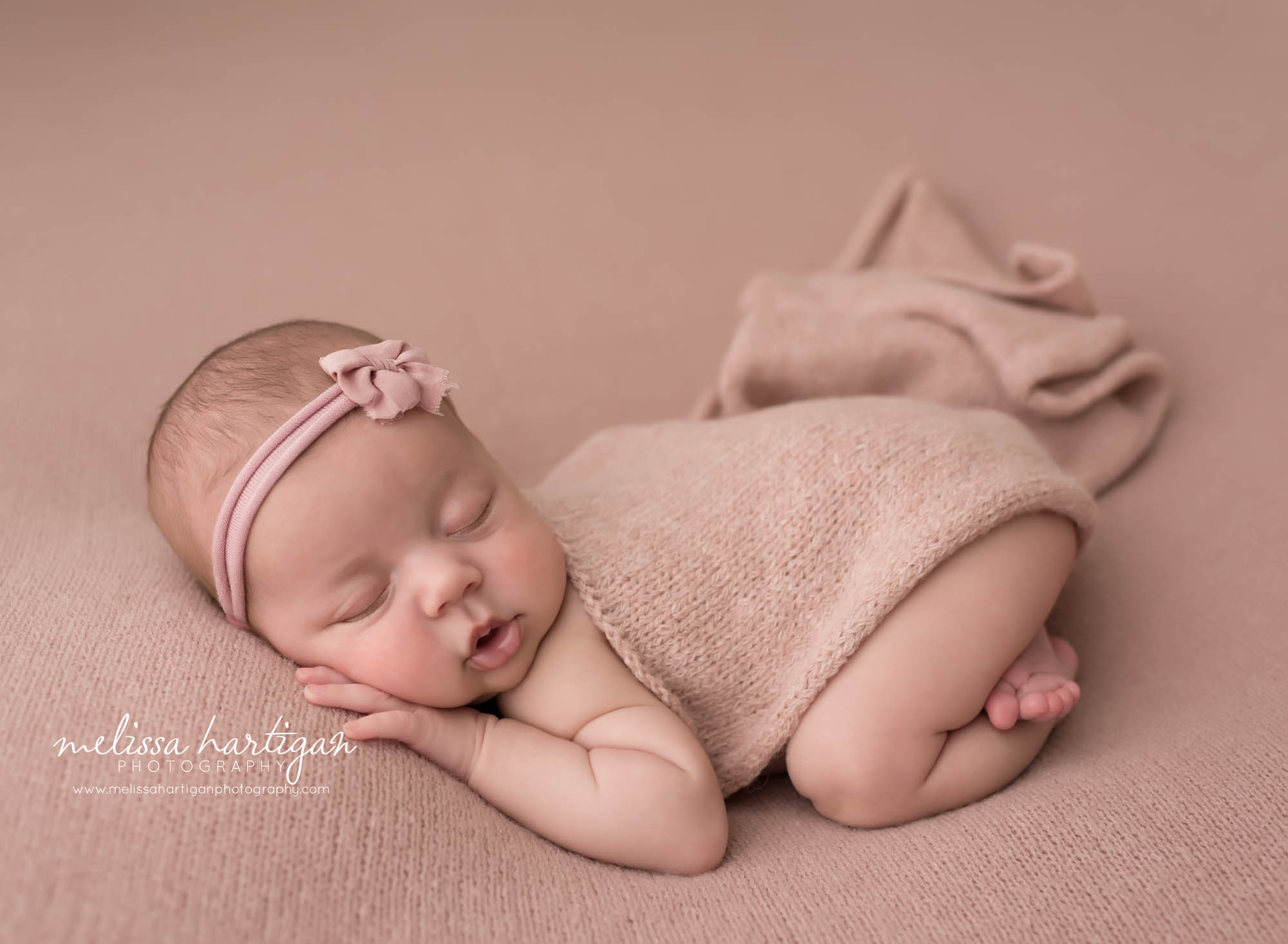 newborn baby girl posed on rose pink colored backdrop wearing pink bow headband and knitted layer draped over her back Newborn photography stafford CT