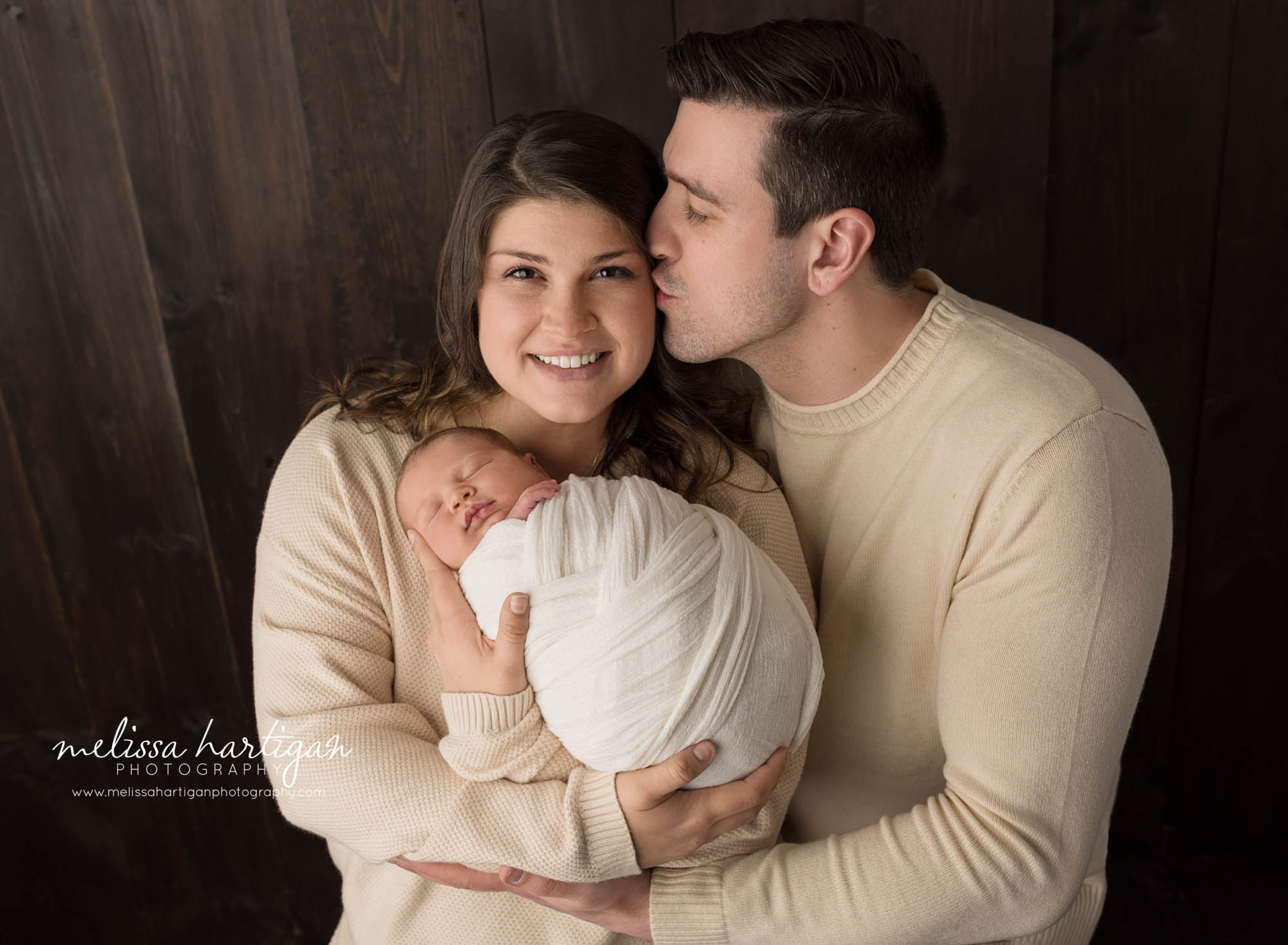 dad kissing mom on side of head mom holding newborn son wrapped in cream white wrap parent pose