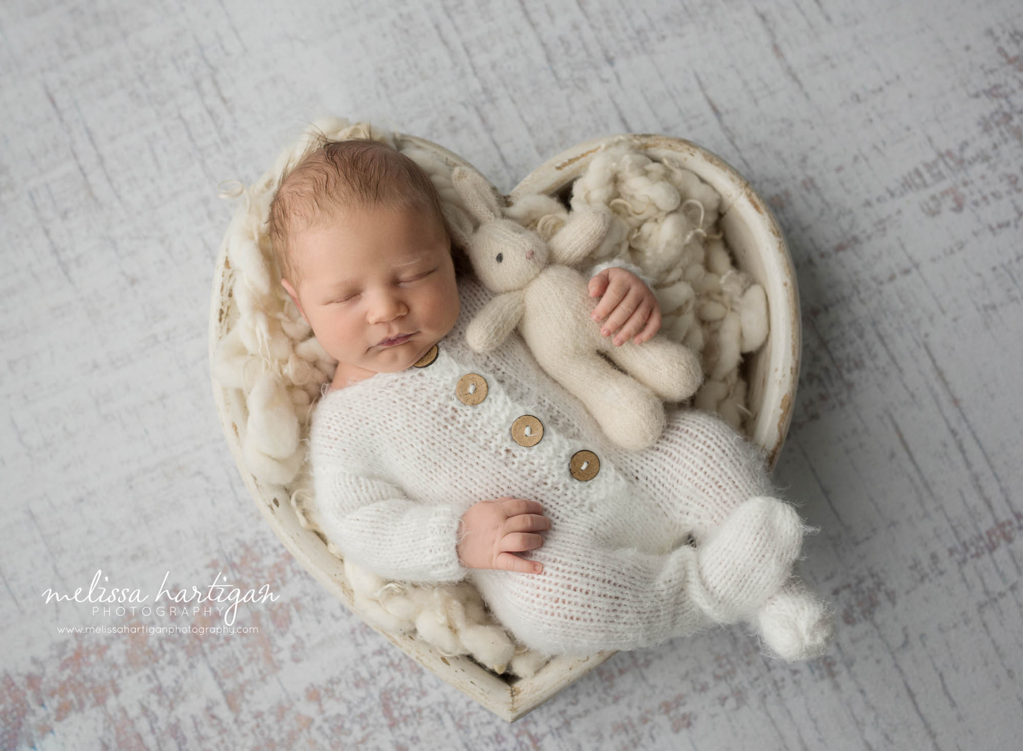 newborn baby boy posed in wooden heart bowl wearing knitted footed outfit holding knitted teddy bear newborn photography windham county