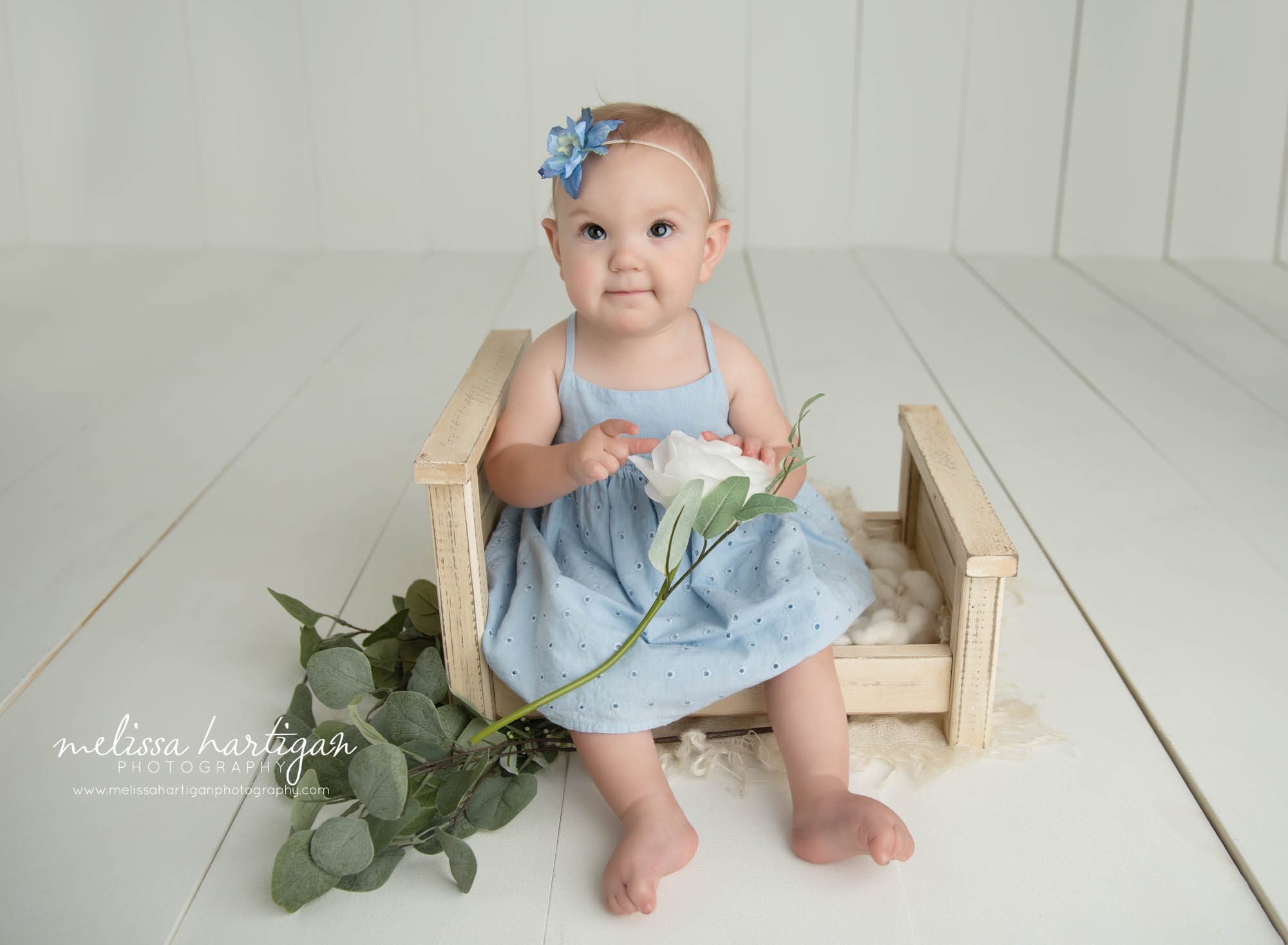 baby girl wearing light blue baby dress sitting on wooden bed prop holding white flower storrs mansfield baby photography