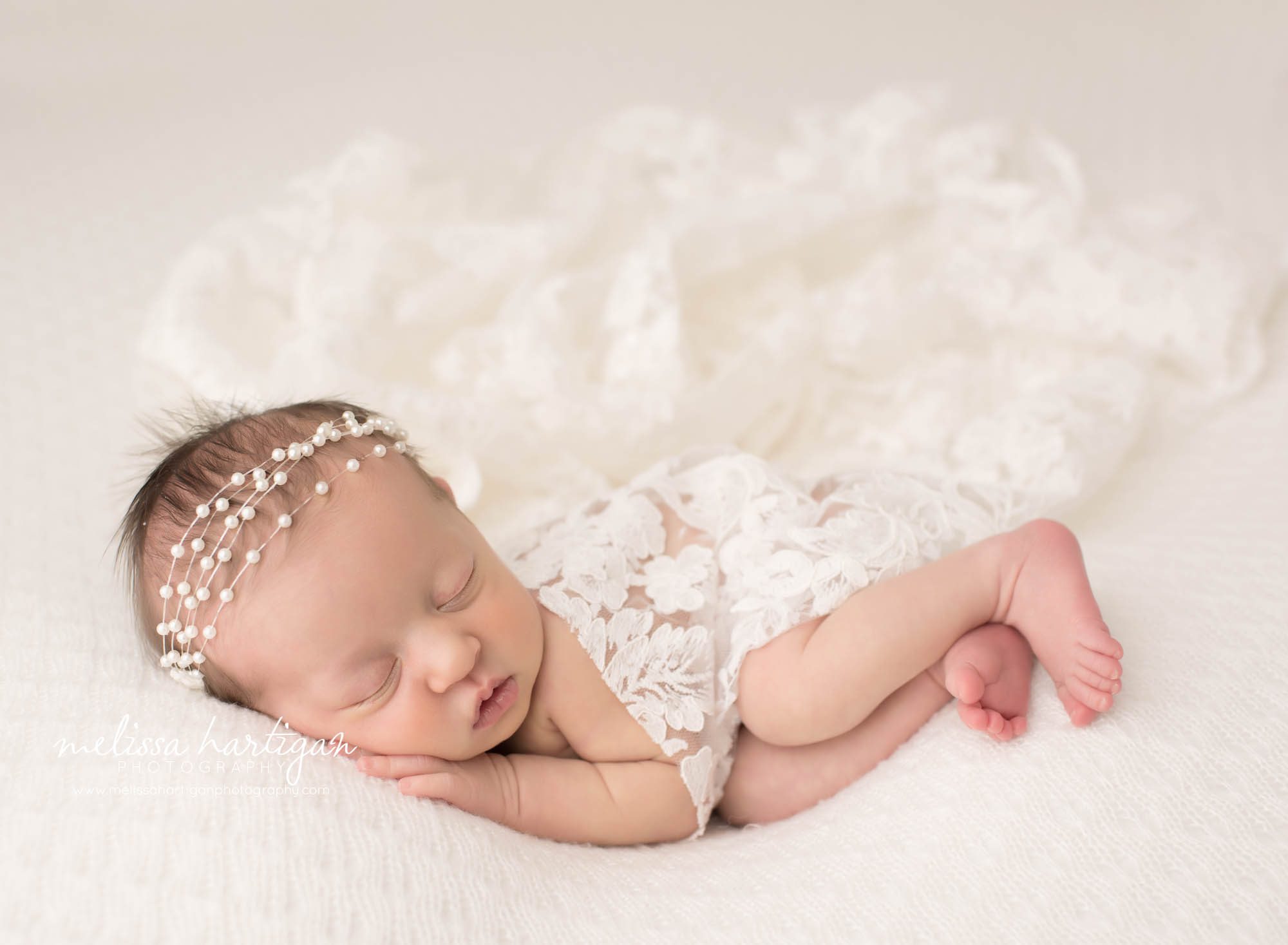 newborn baby girl posed on cream backdrop with white lace layer draped over her posed on side