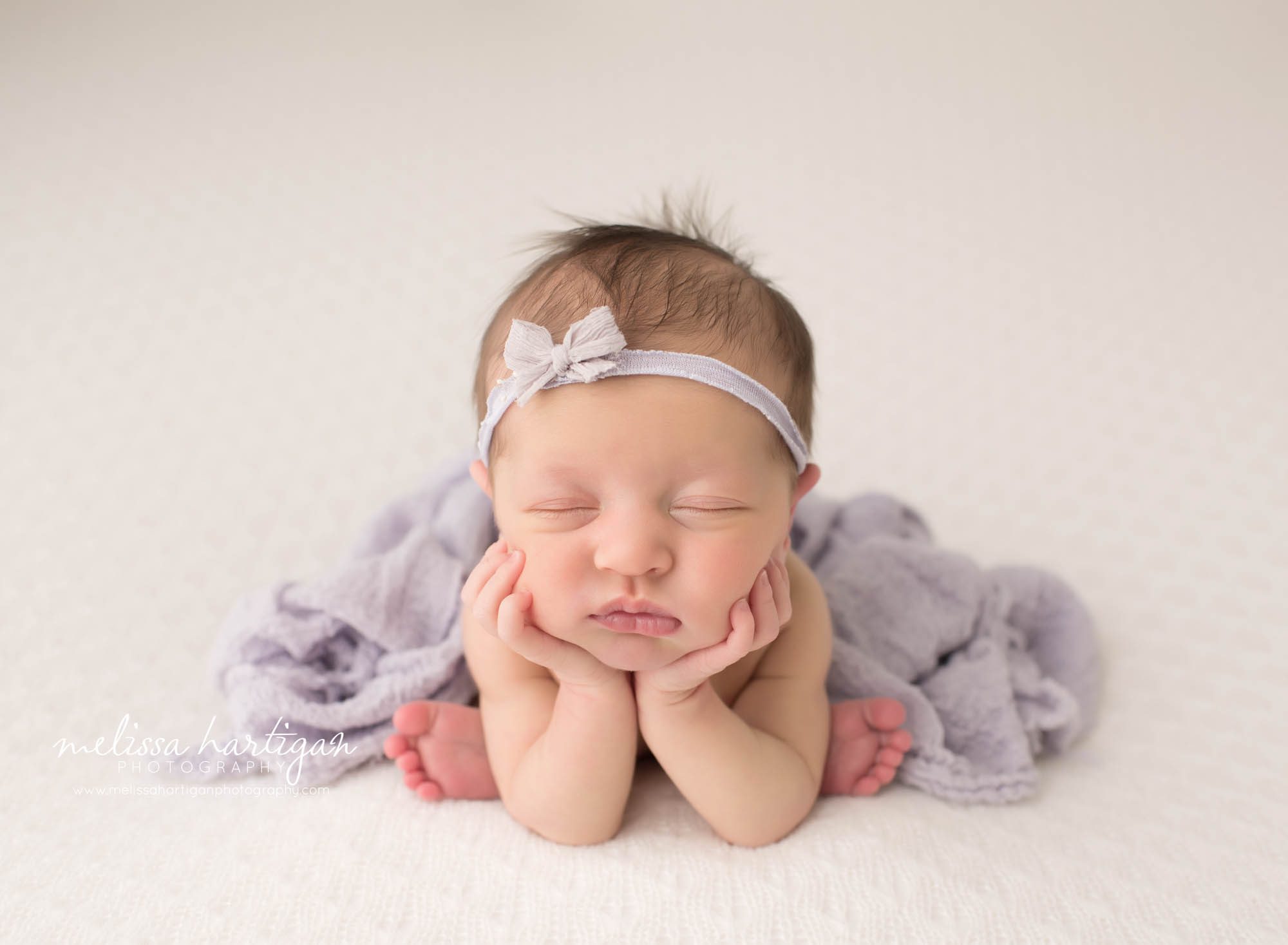 newborn baby girl posed on cream backdrop in froggy pose wearing purple bow headband and purple knitted layer middlesex county newborn photography
