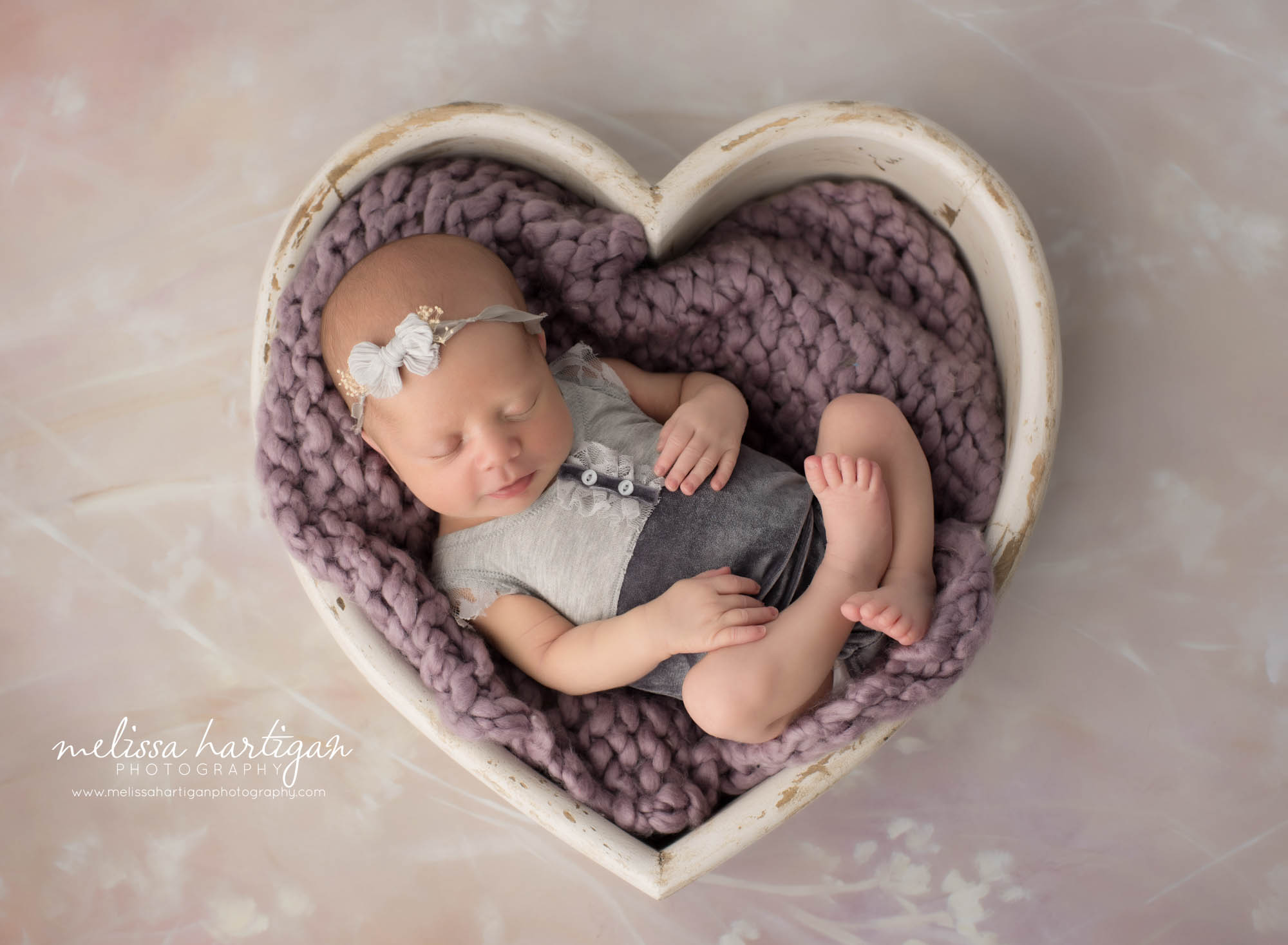 newborn baby girl posed in cream wooden heart bowl prop wearing grey newborn baby outfit and bow headband
