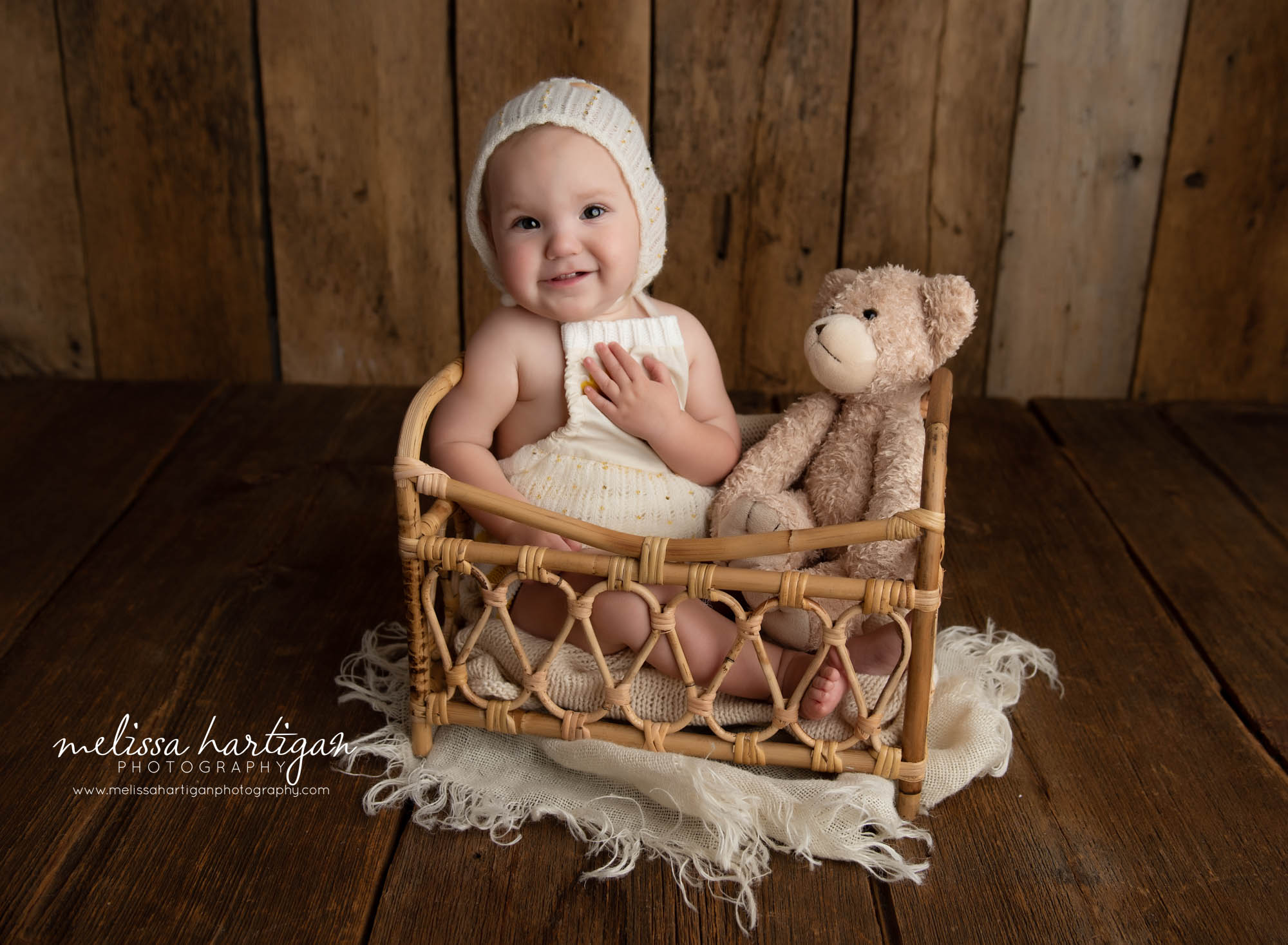 baby girl sitting in basket wearing knitted baby bonnet with teddy bear holding hand to heart
