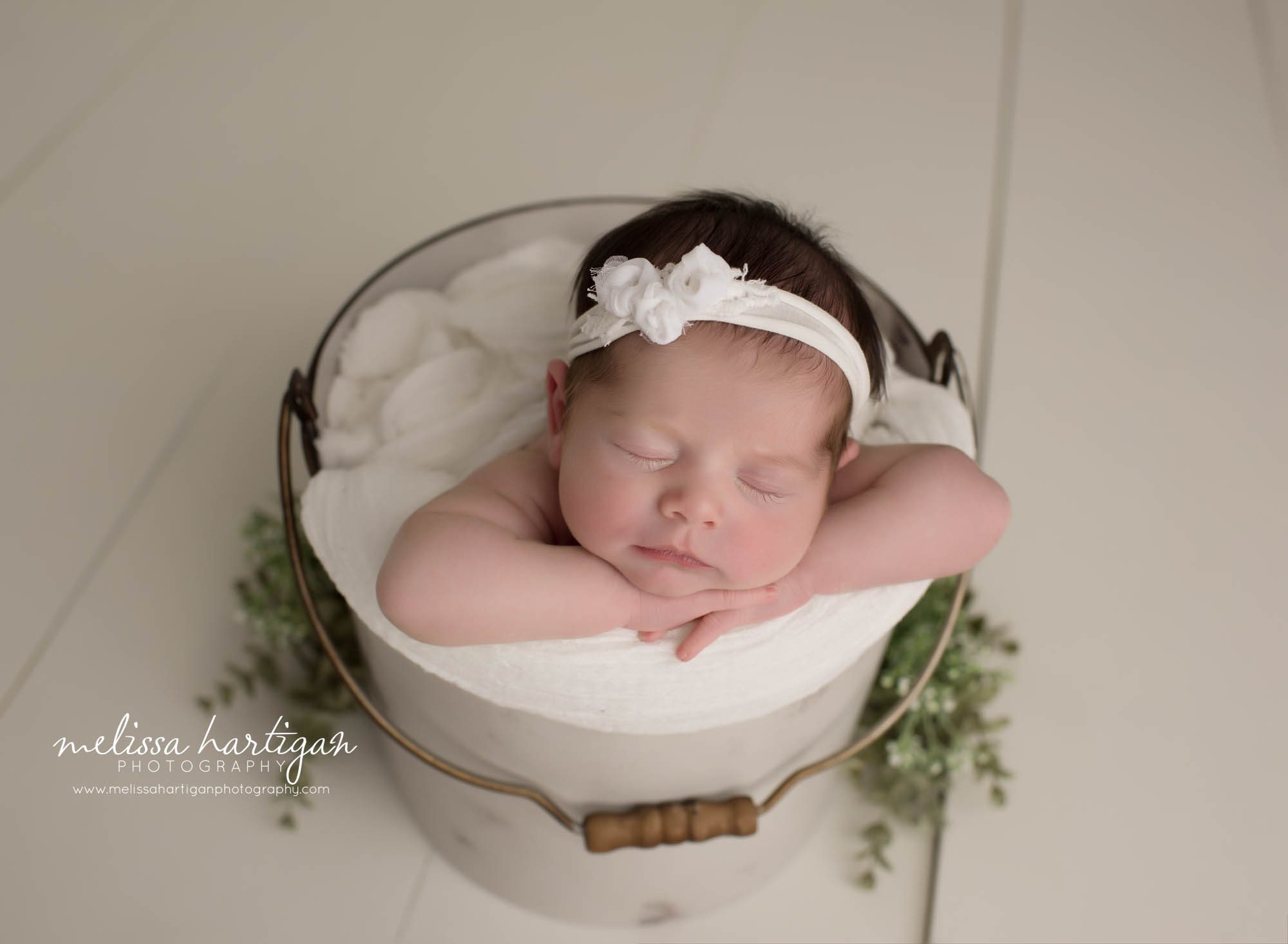 newborn baby girl posed in bucket wearing white bow headband