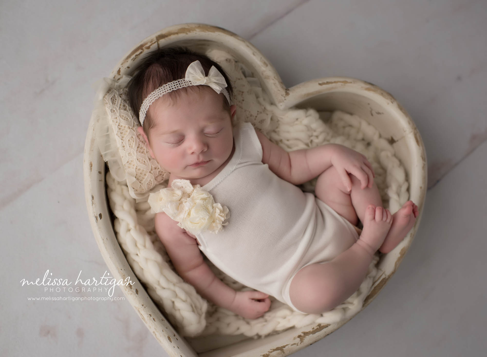 newborn baby girl posed in wooden heart bowl prop wearing white newborn outfit and bow headband colchester CT newborn photography