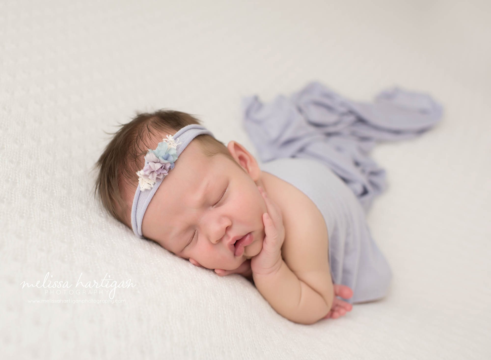 newborn baby girl posed on cream backdrop wearing purple flower headband and purple draping wrap