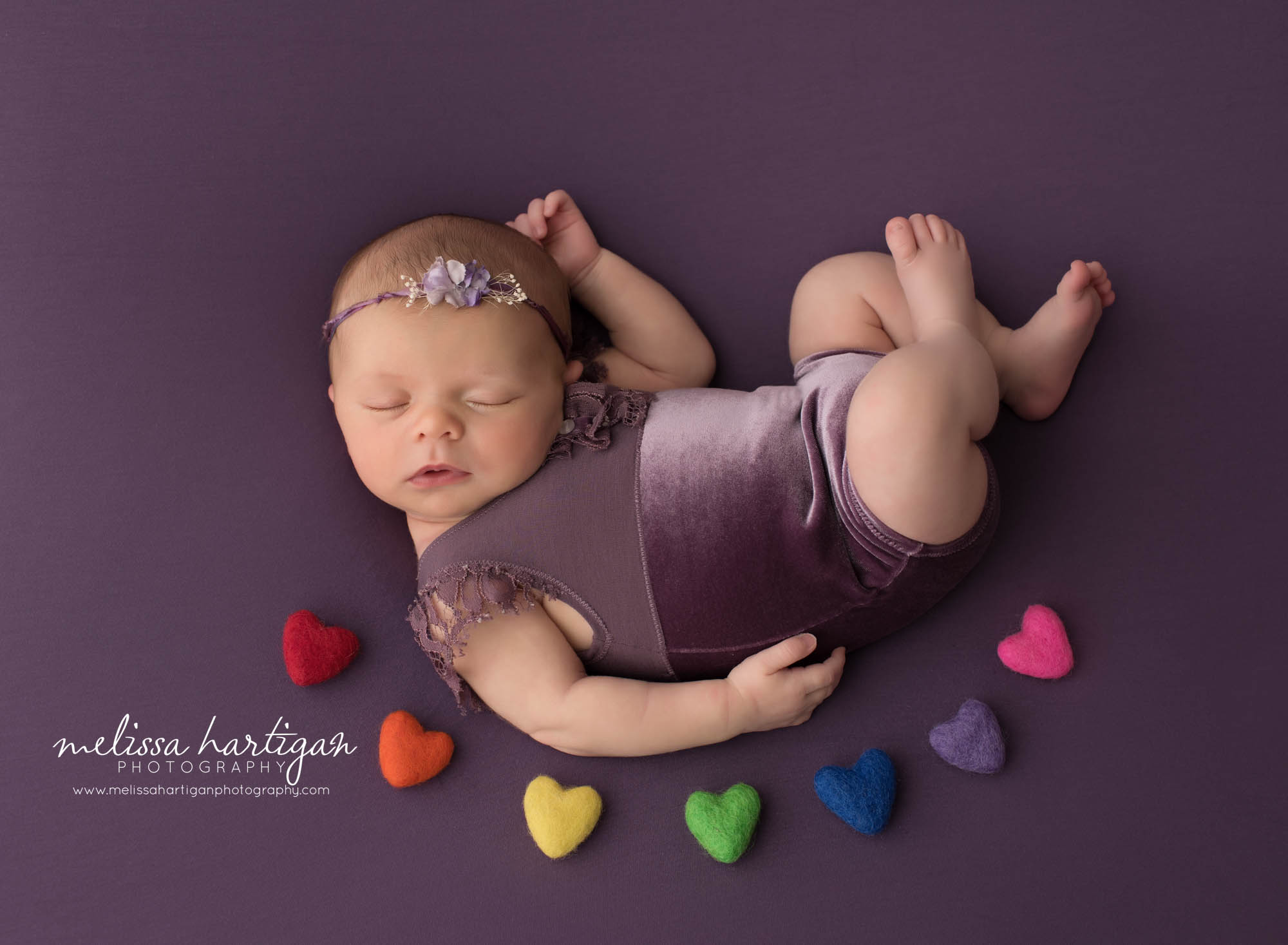 newborn baby girl posed on purple backdrop wearing purple outfit and rainbow felted hearts