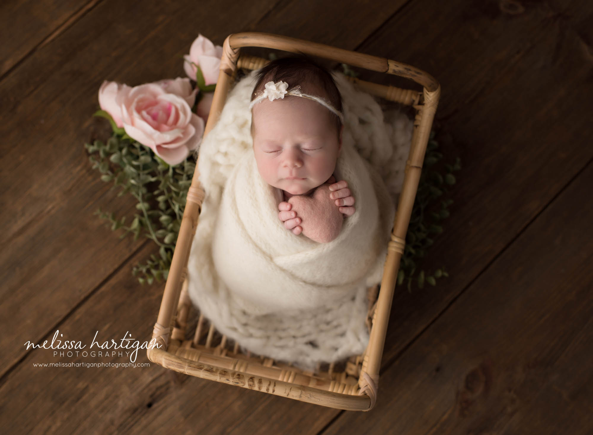 newborn baby girl wrapped in cream knit wrap posed in basket with pink flowers beside basket