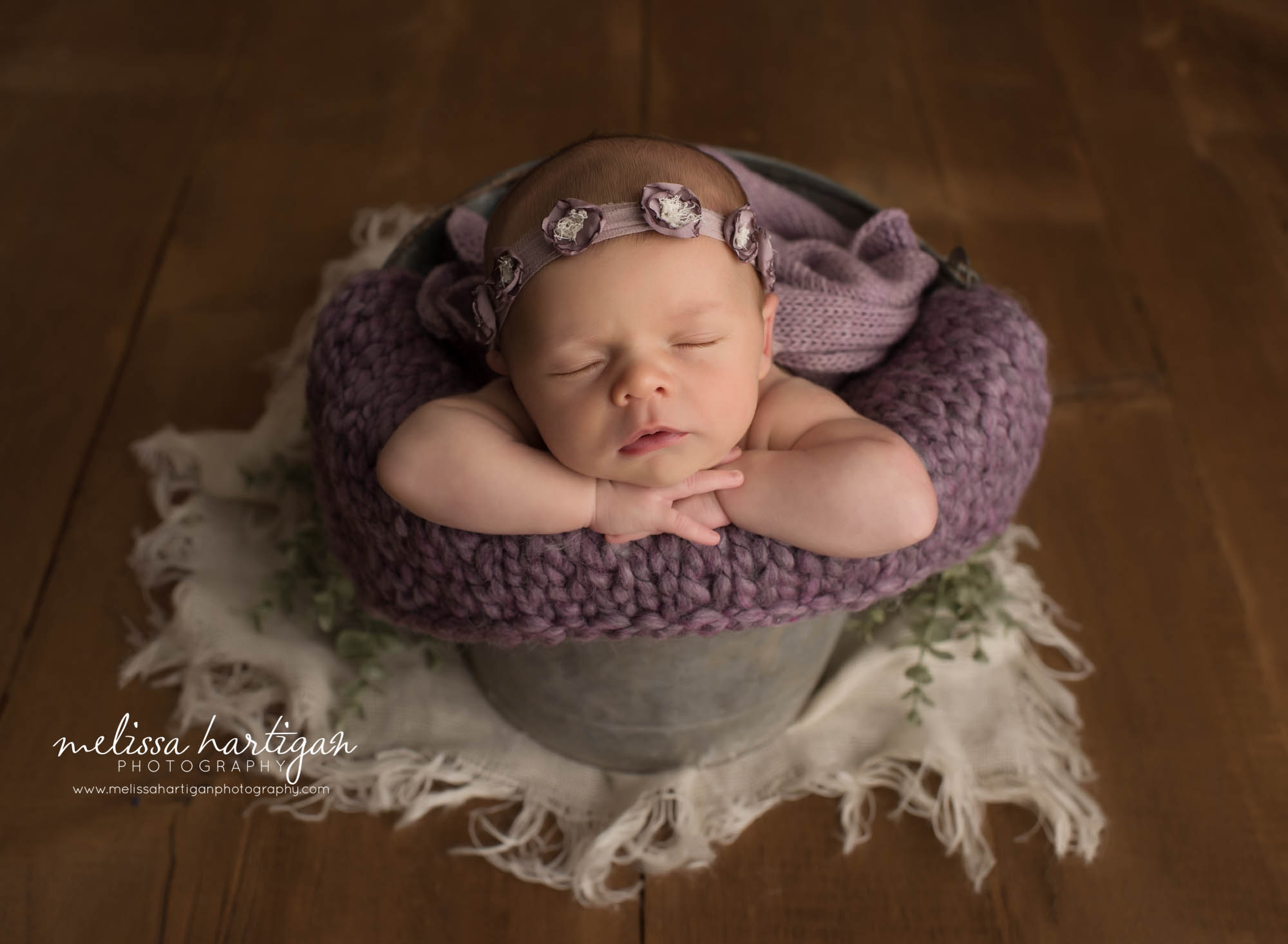 newborn baby girl posed in bucket with purple knitted layer wearing purple flower headband
