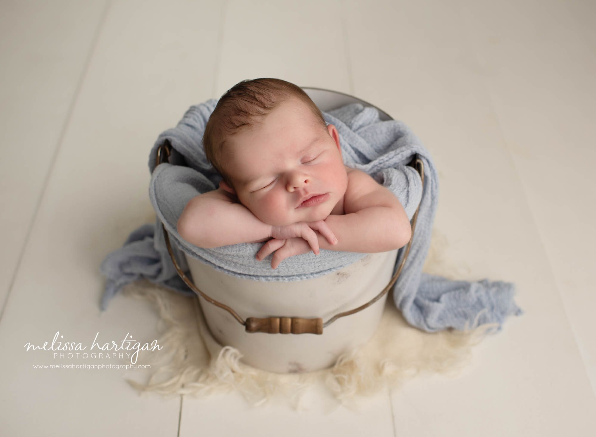 newborn baby boy posed in cream bucket with blue layer wrap draped over his back