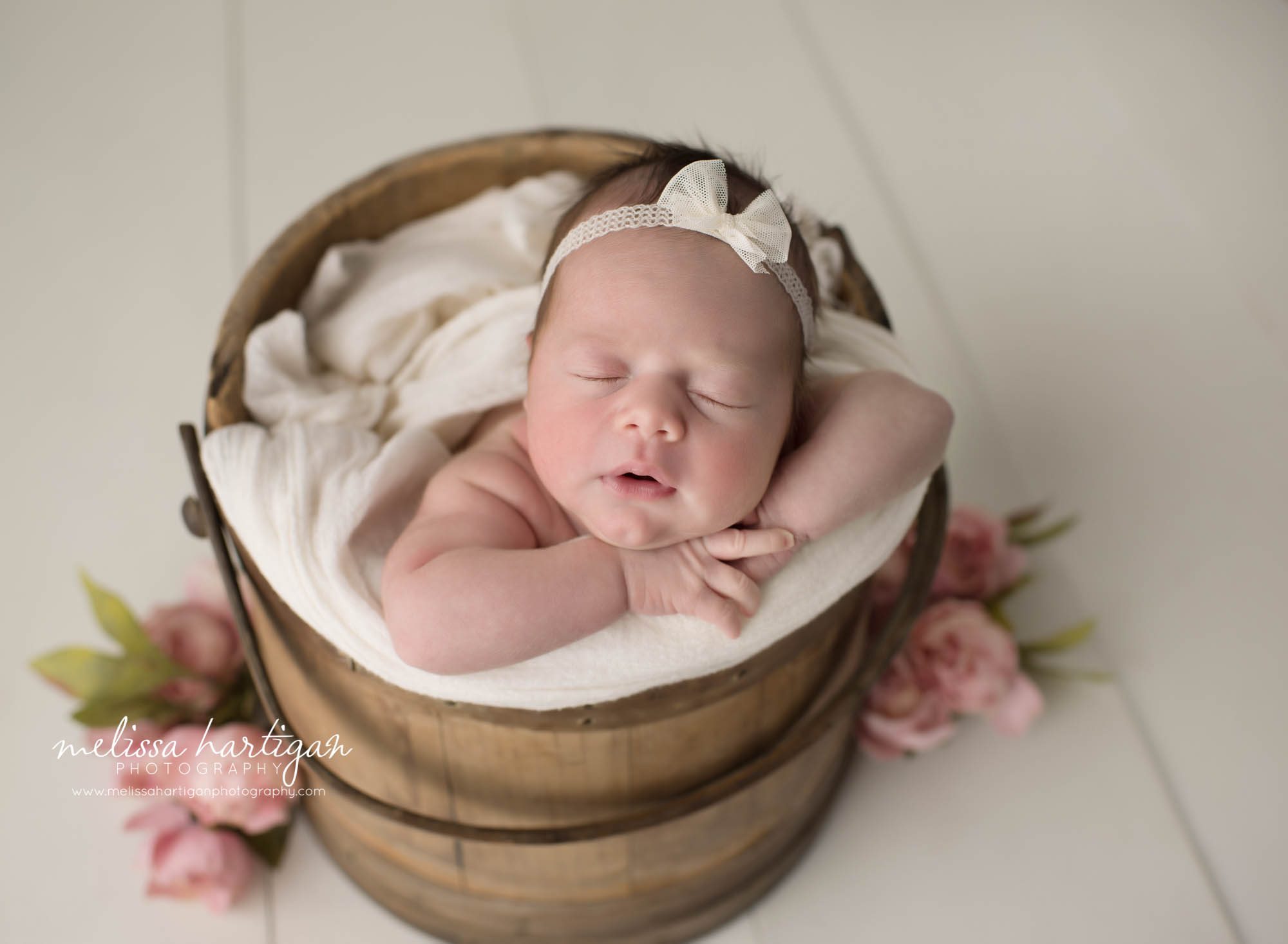 newborn baby girl posed in wooden barrell wearing white bow headband