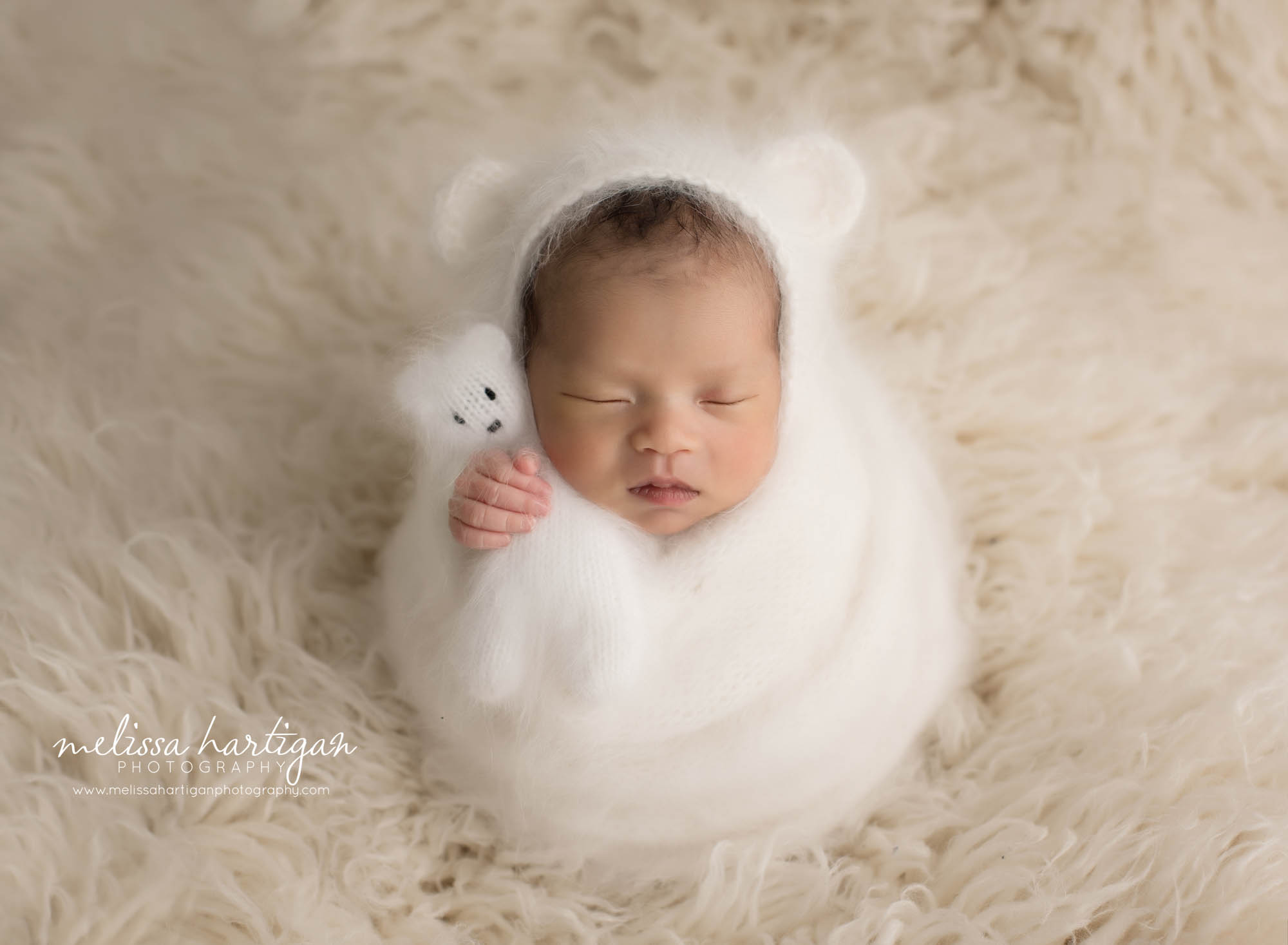 baby girl wrapped in white knitted layer wrap wearing matching bear bonnet and holdinng teddy bear lovey