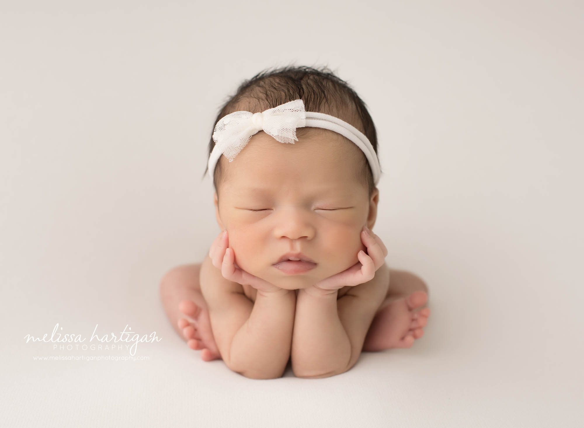 newborn baby girl posed froggy pose on white backdrop wearing white bow headband
