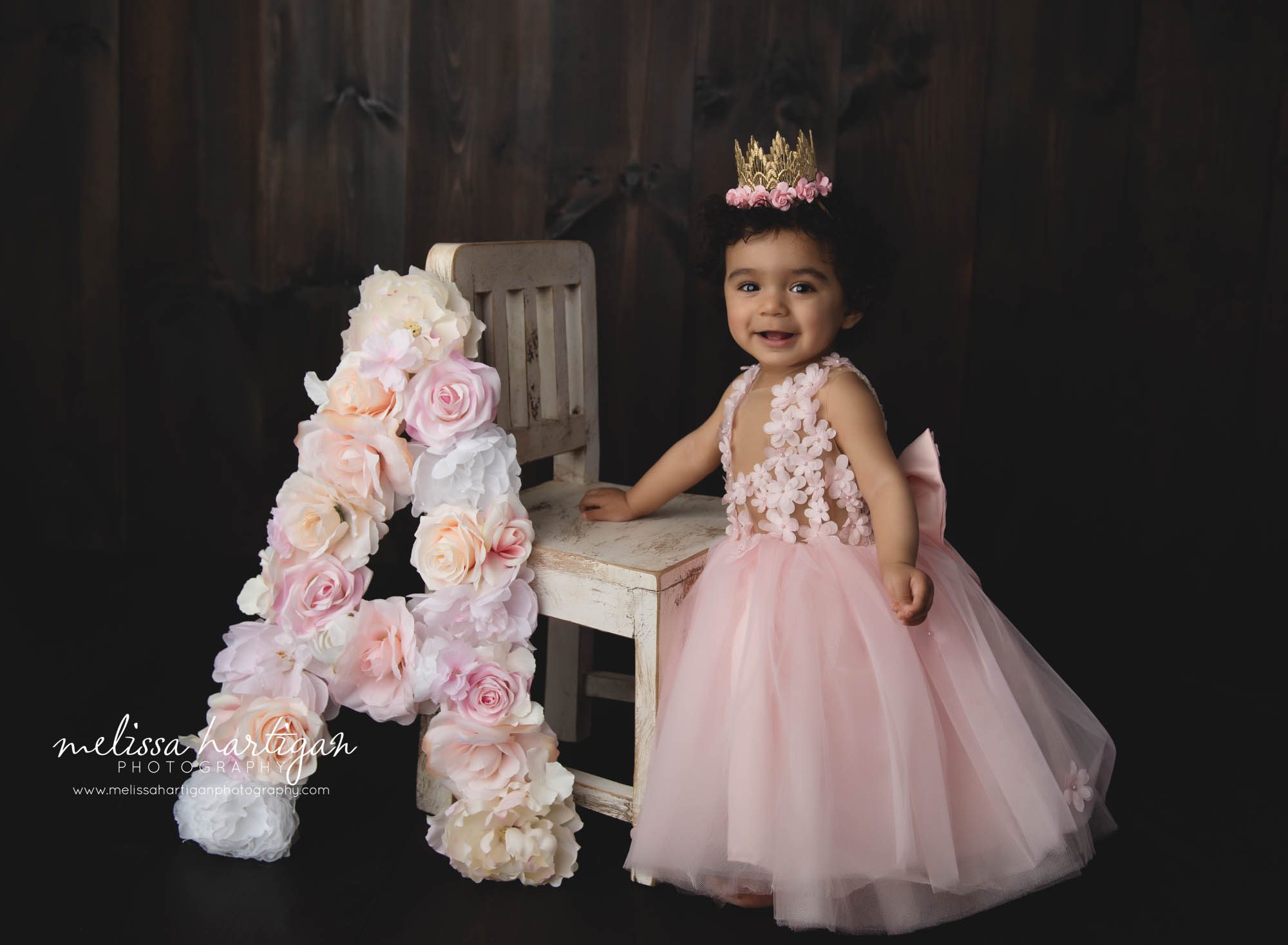 baby girl standing next to wooden chair with floral A letter wearing pink dress and crown