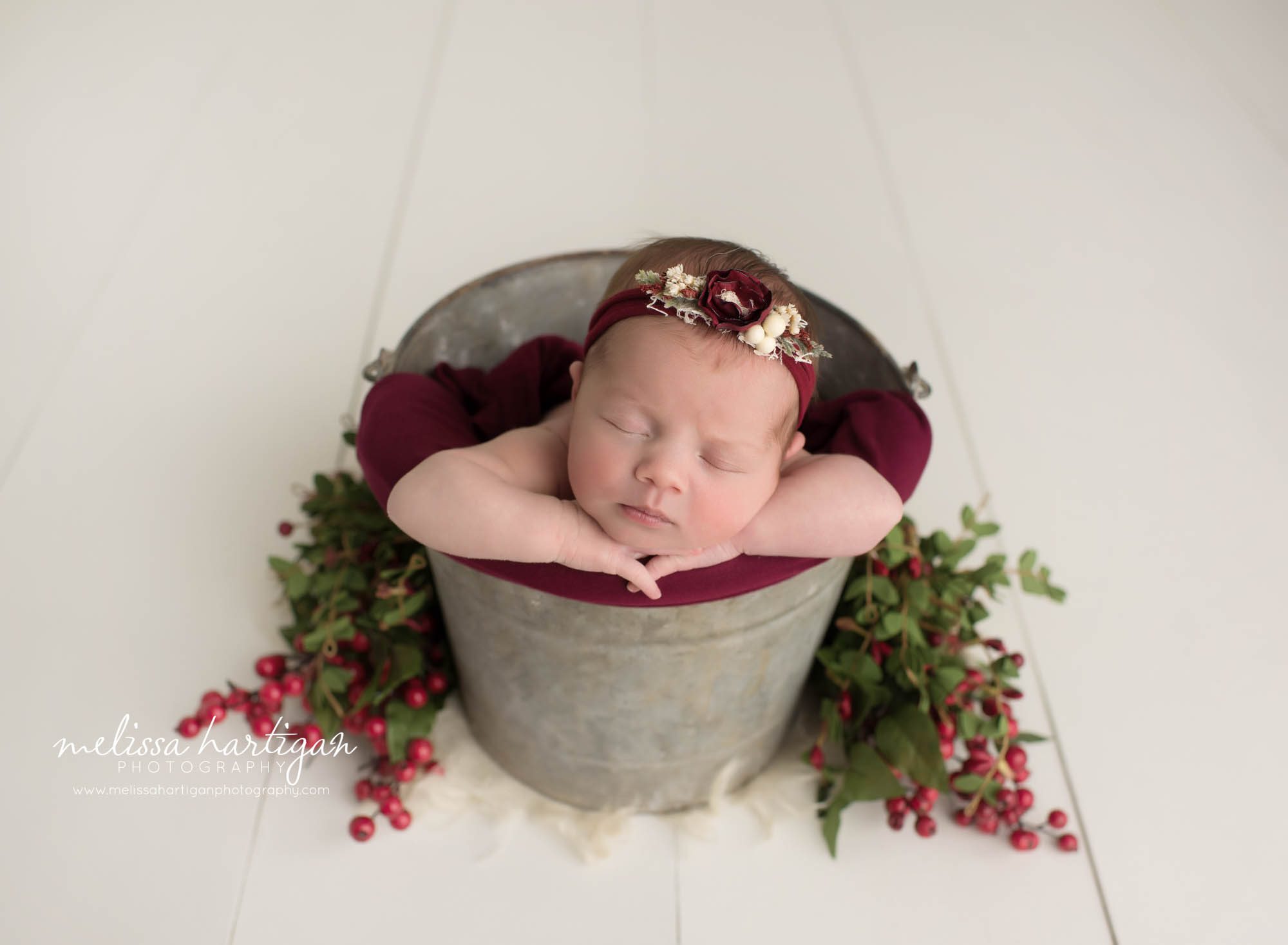 newborn baby girl posed in bucket wearing red headband with red and green foilage elements