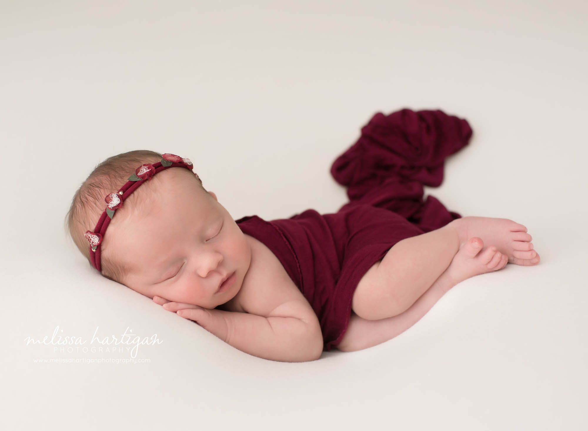 newborn baby girl posed on side on white backdrop wearing red flower headband red drape layer