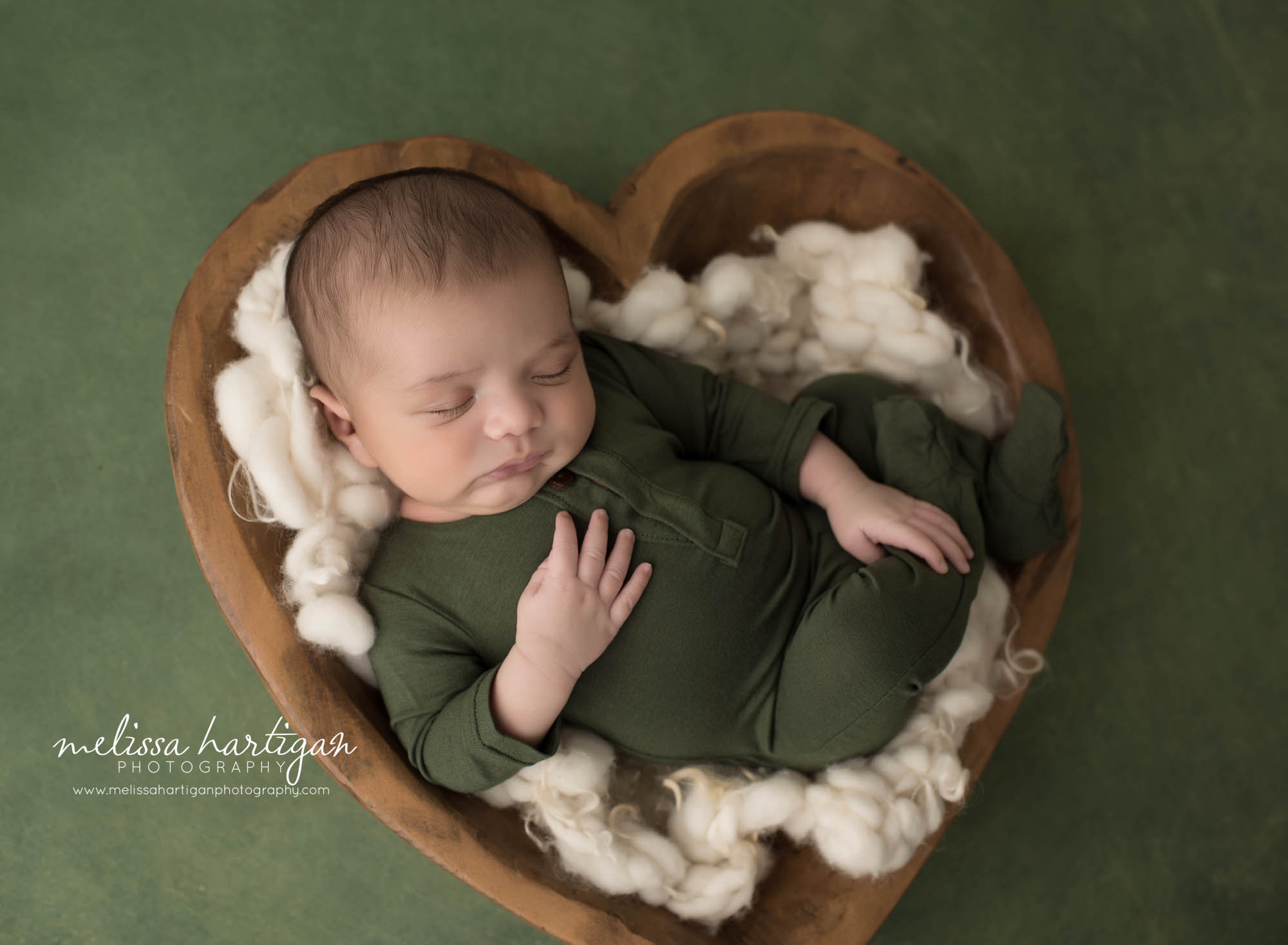 newborn baby boy wearing green footed romper posed in wooden heart bowl prop with cream fluffy layer