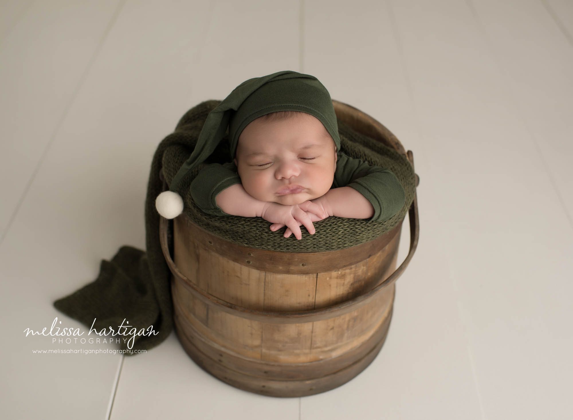 newborn baby boy posed on wooden barrel wearing green sleepy cap with cream pom on it