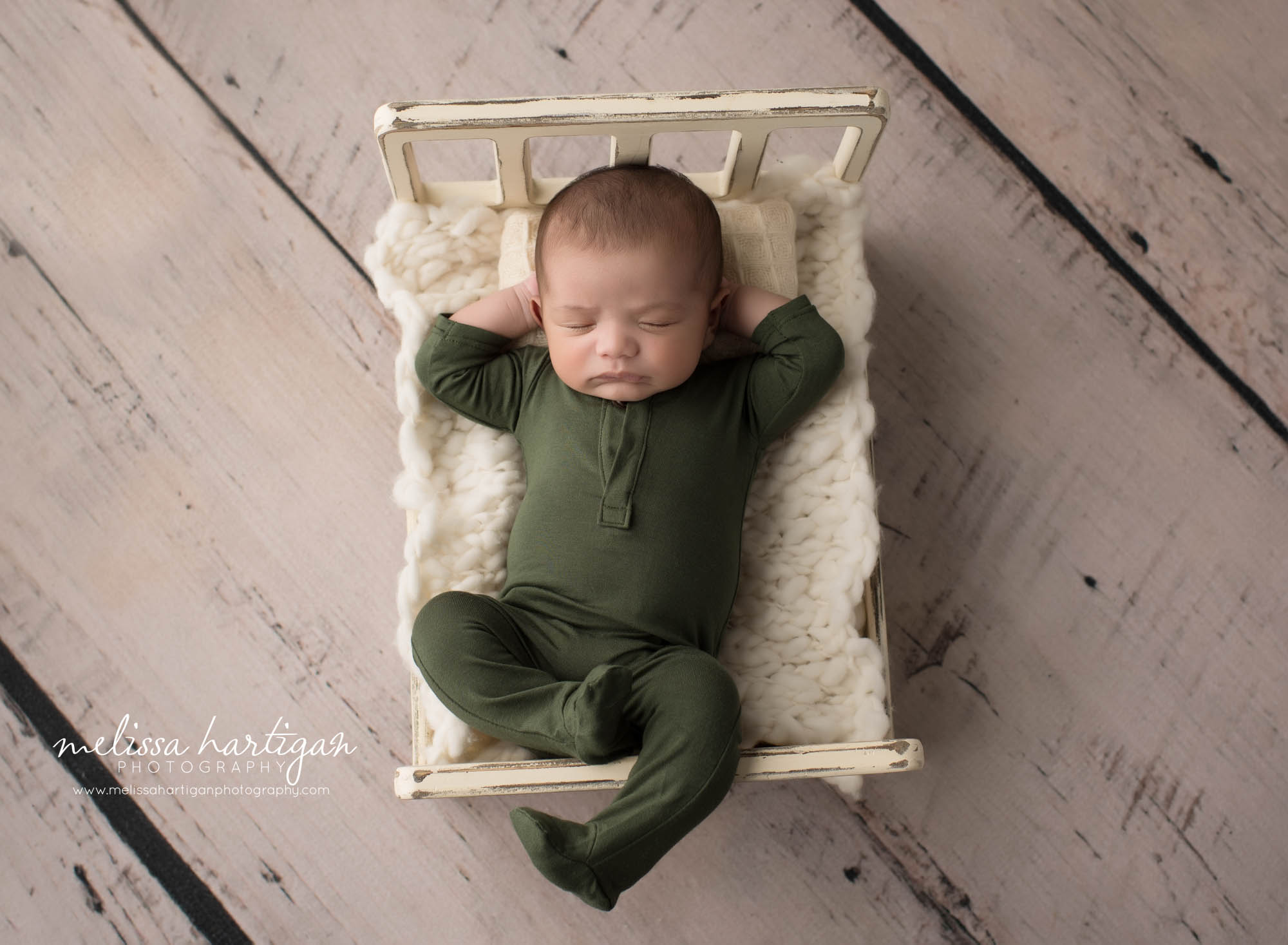 newborn baby boy wearing green footed romper posed on back in wooden bed prop with hands resting under back of his head connecticut newborn photography