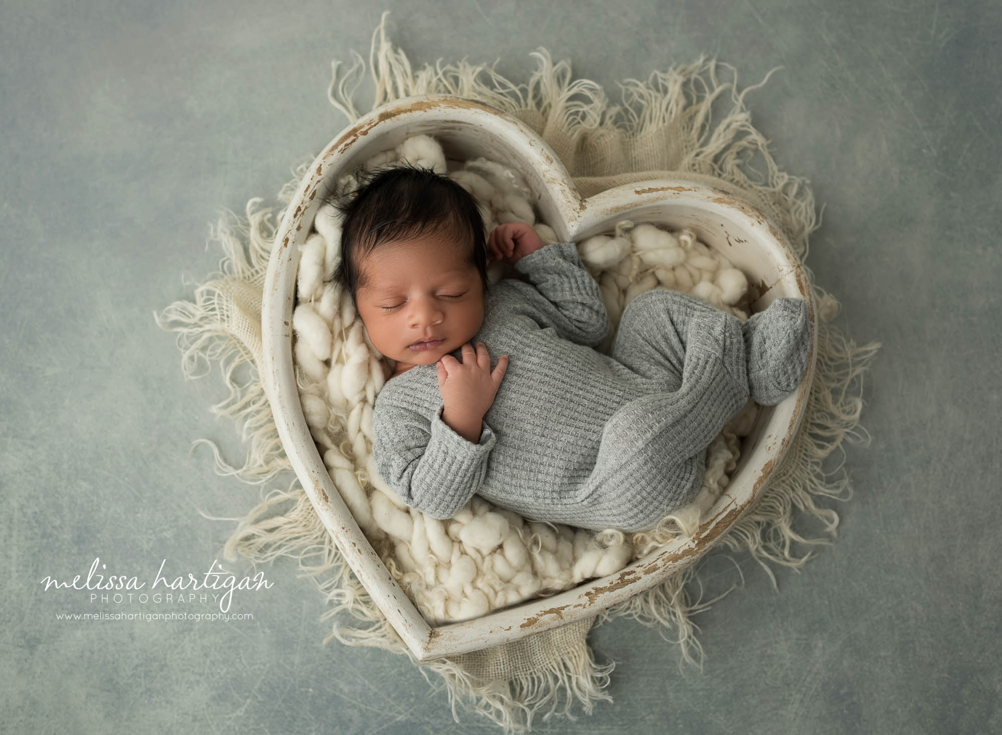 newborn baby boy wearing gray footed baby sleeper posed in cream wooden heart bowl prop