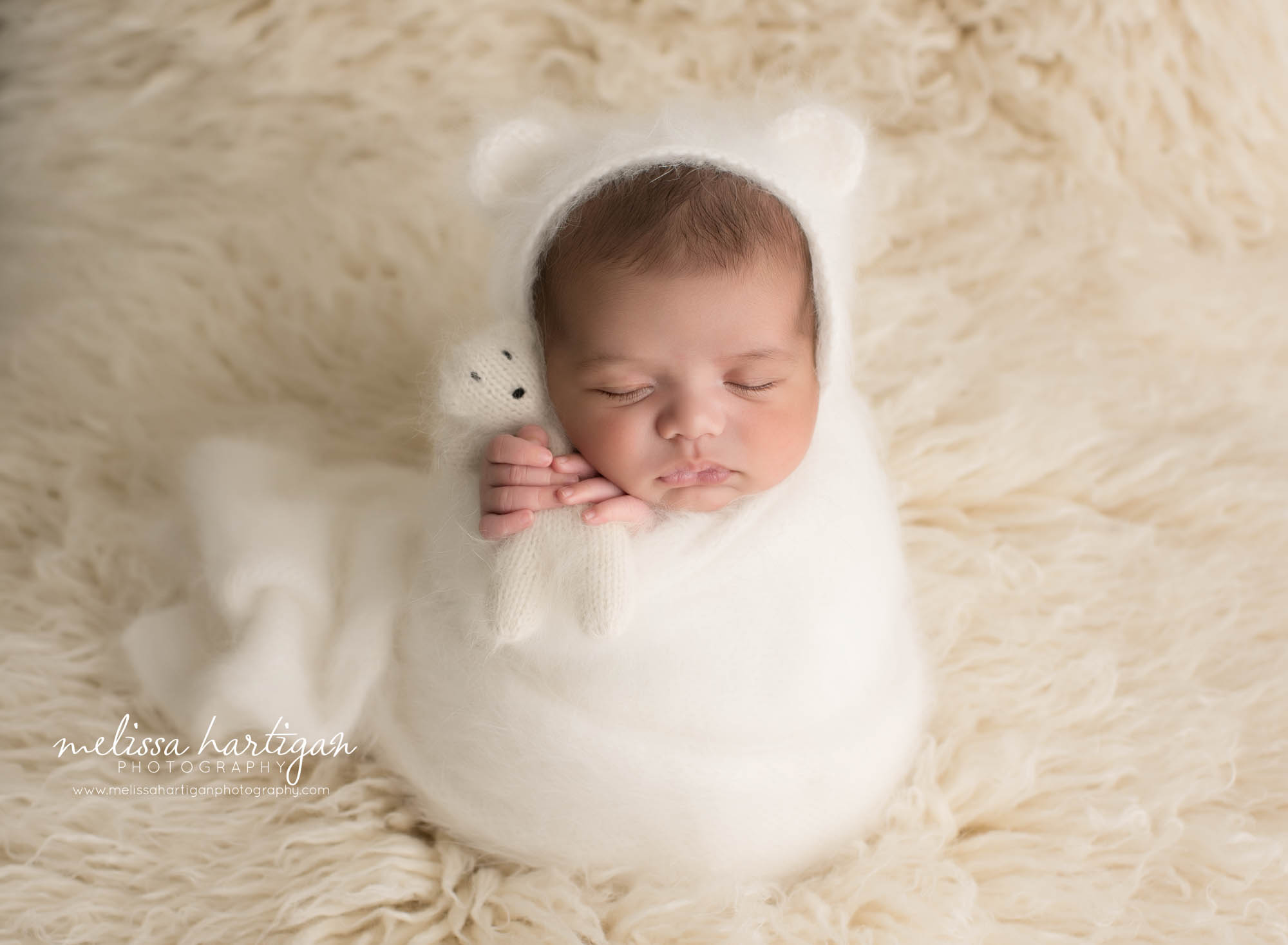 newborn baby boy wrapped inc ream knitted wrap holding knitted teddy bear and wearing matching knitted bear bonnet