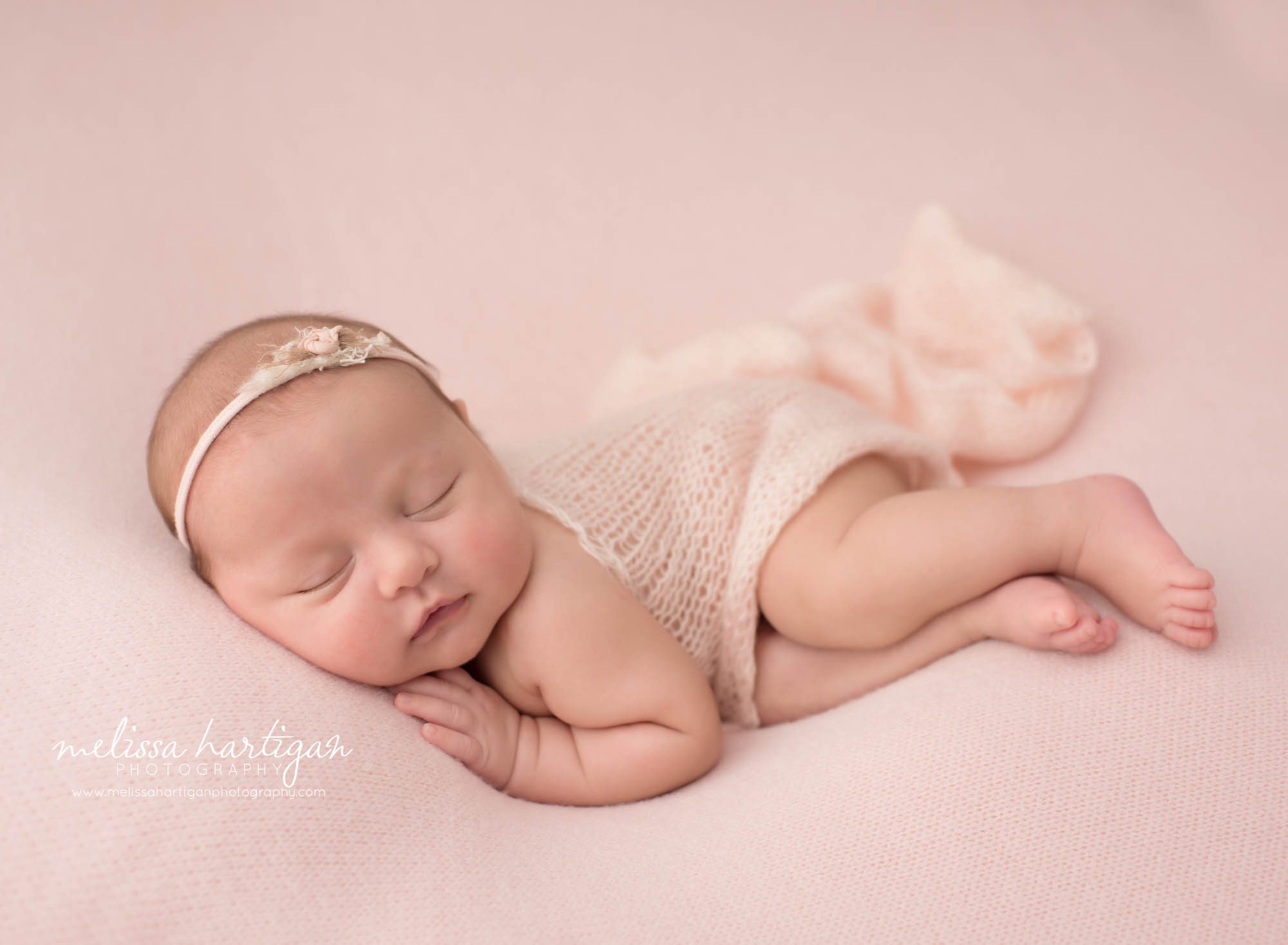 newborn baby girl posed on side wearing flower headband and pink knit layer draped over back