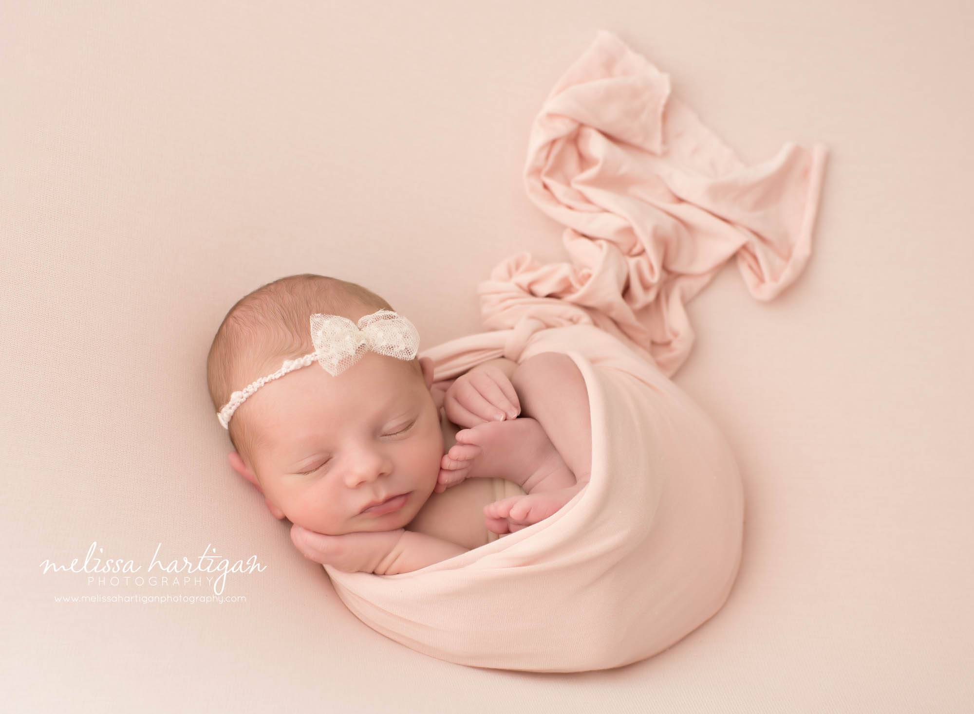 newborn baby girl posed on blush pink backdrop wraped in matching layer wrap wearing cream white bow headband ct new born photography connecticut