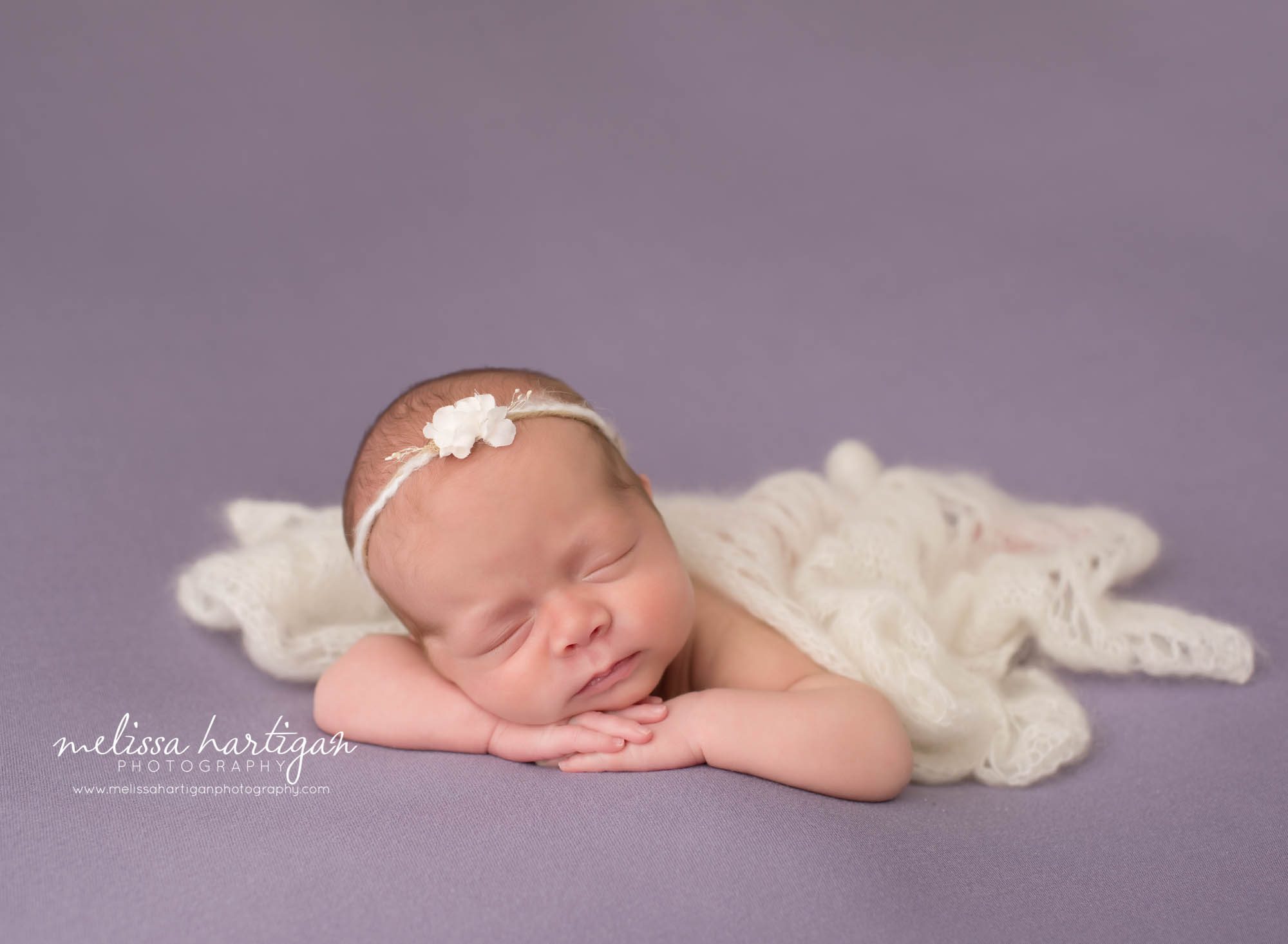 newborn baby girl posed on tummy on purple backdrop with cream drape layer and cream headband