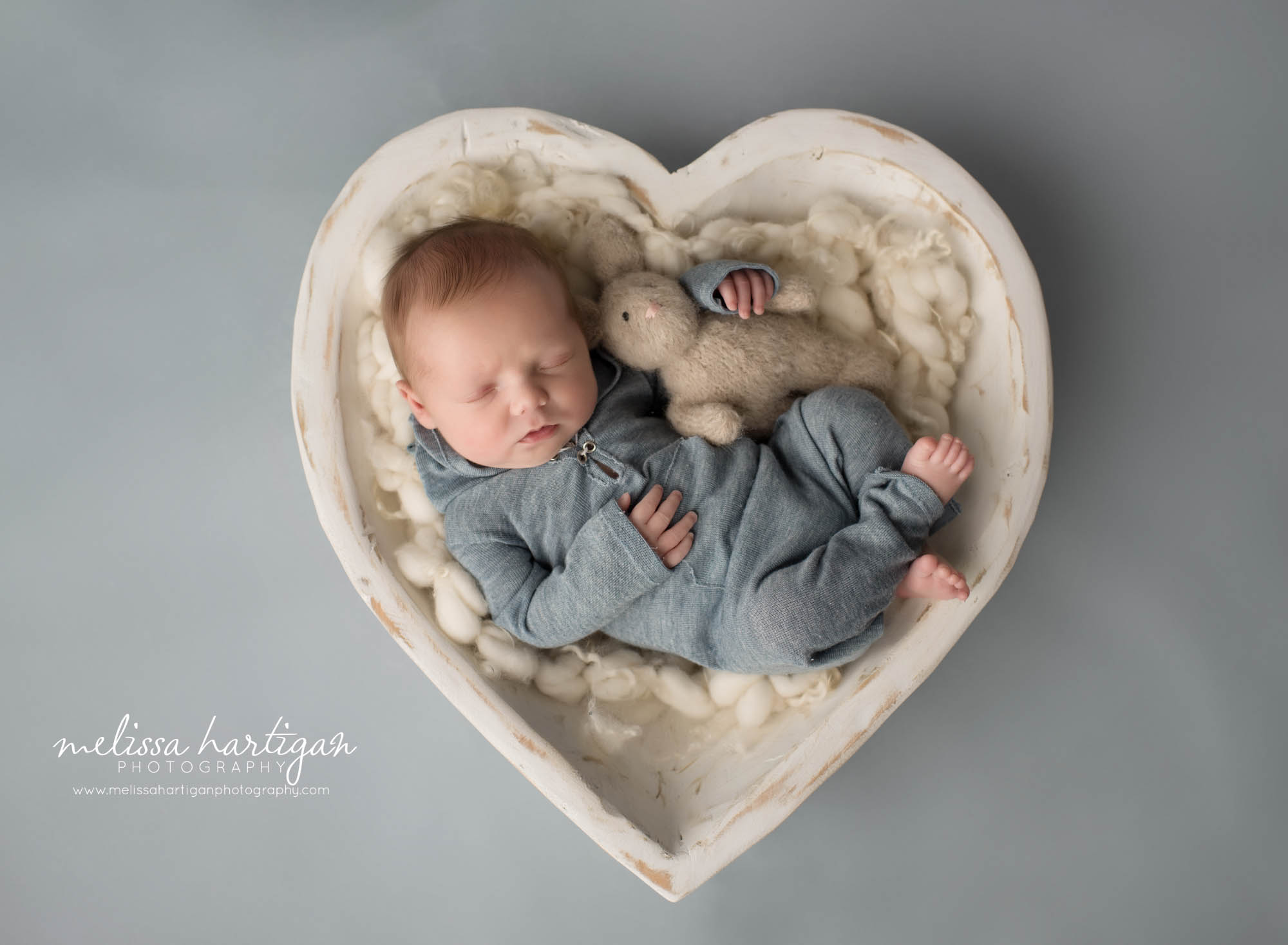newborn baby boy posed in cream wooden heart bowl prop wearing blue outfit holding teddy