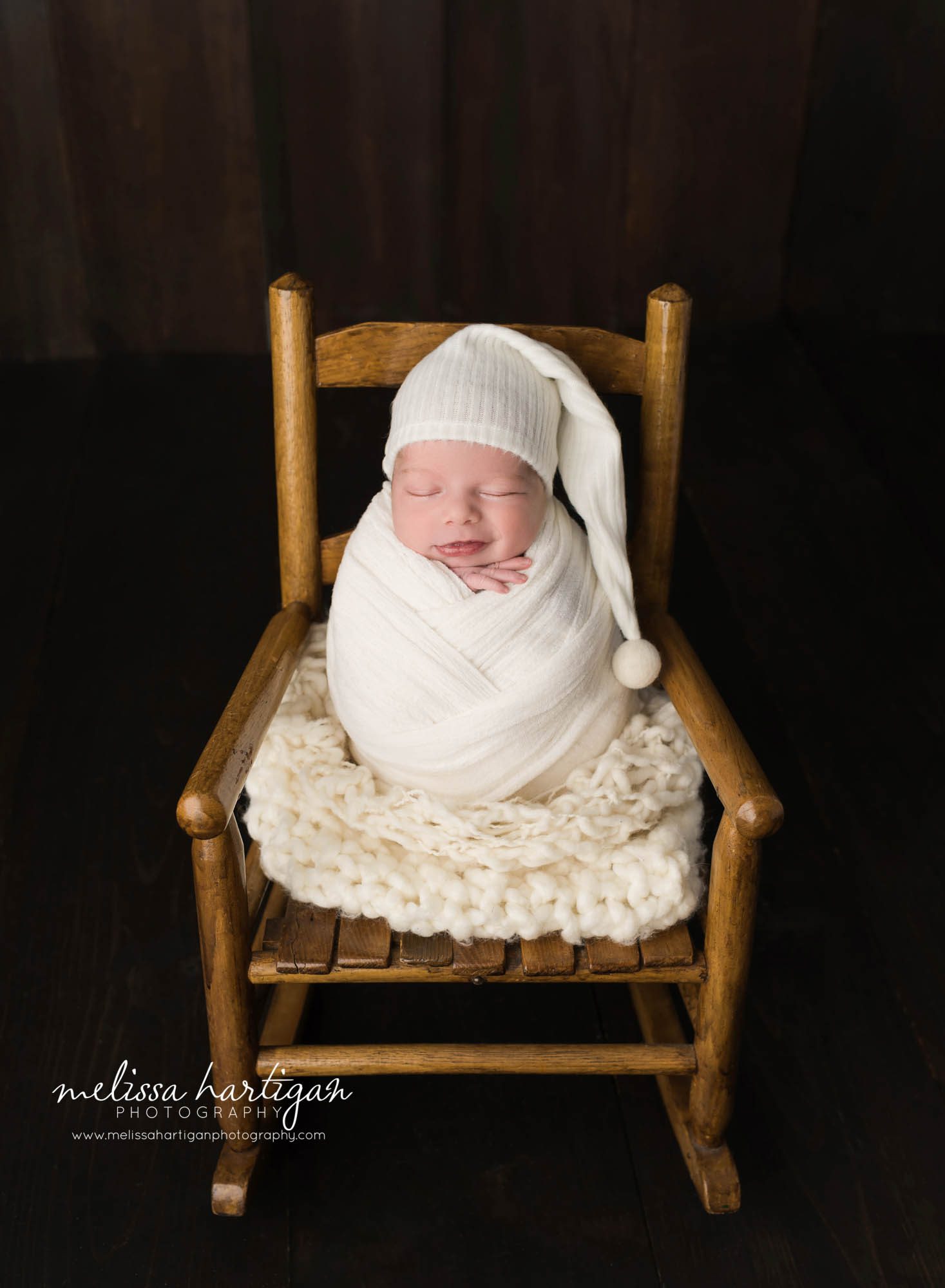 newborn baby boy posed on wooden rocking chair wrapped in cream wrap smiling and wearing cream sleepy cap