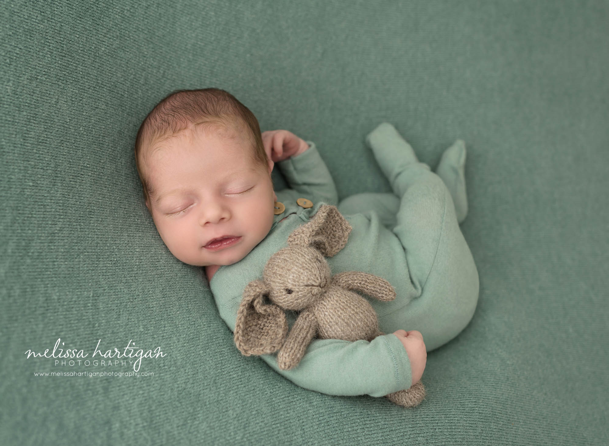 newborn baby boy wearing green sleeper outfit pose don green backdrop holding bunny lovey