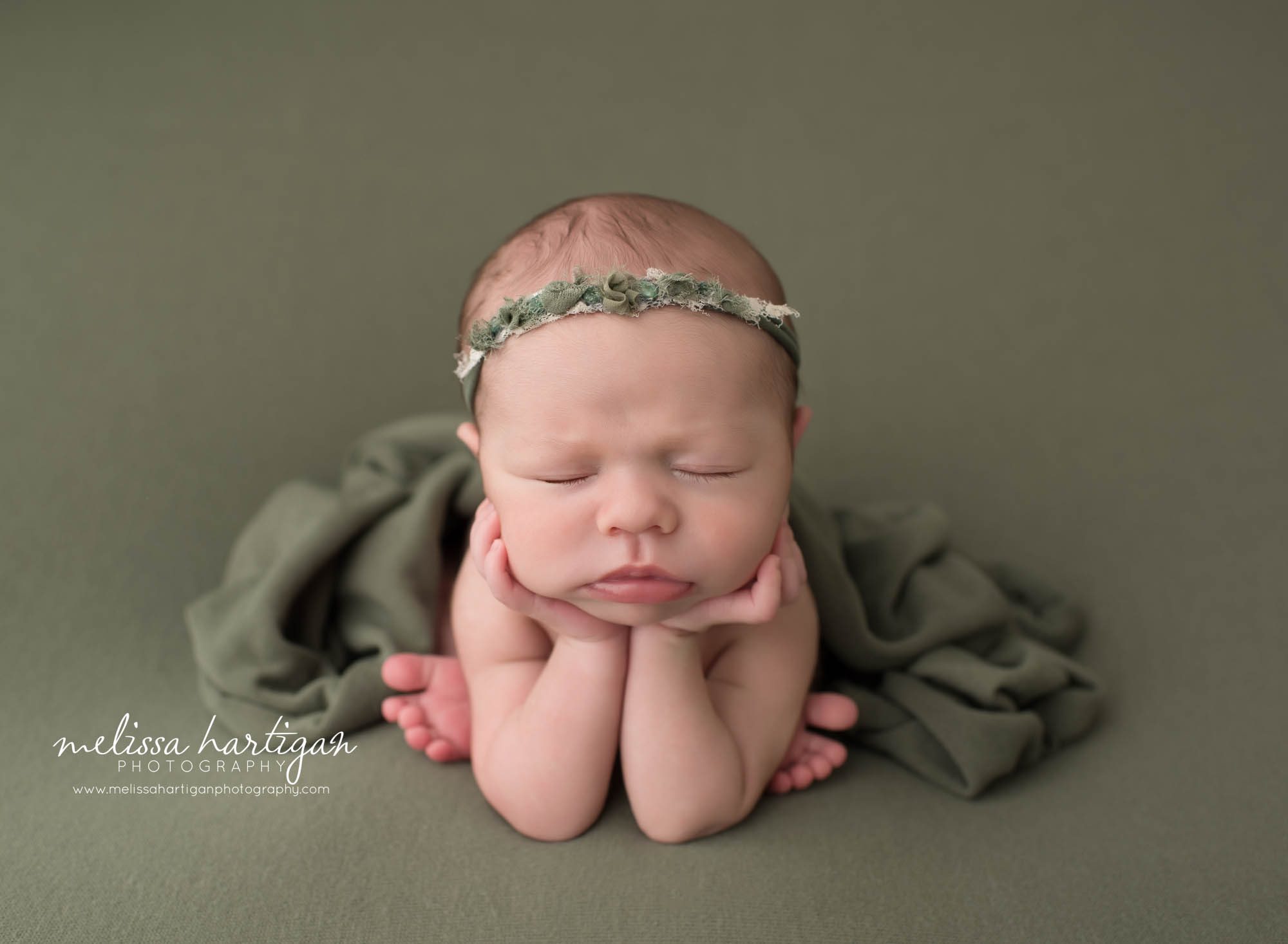 newborn baby girl posed in froggy pose on olive green colored backdrop wearing headband