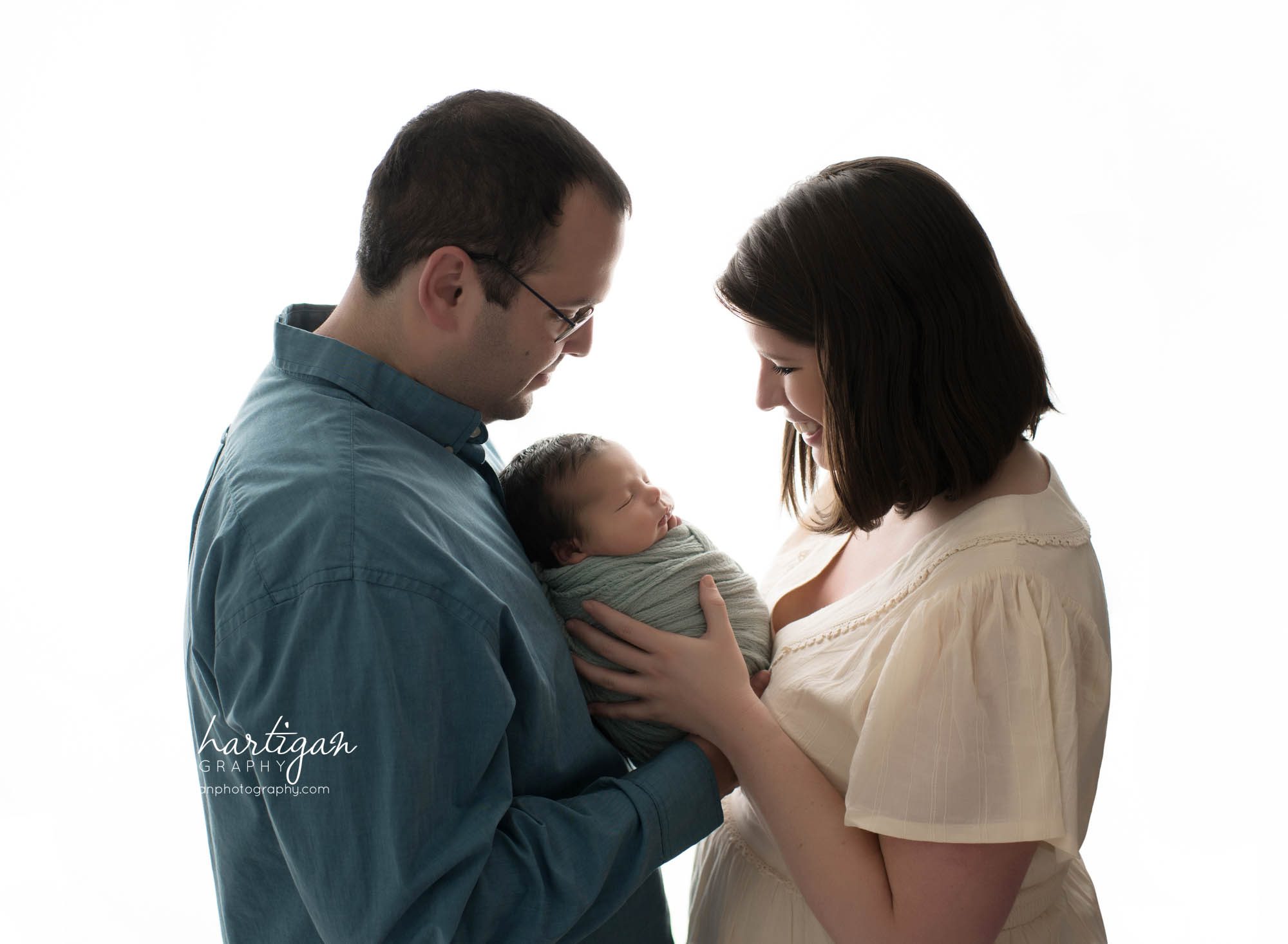 newborn baby boy wrapped in soft teal colored wrap posed with mom and dad facing each other parents family pose