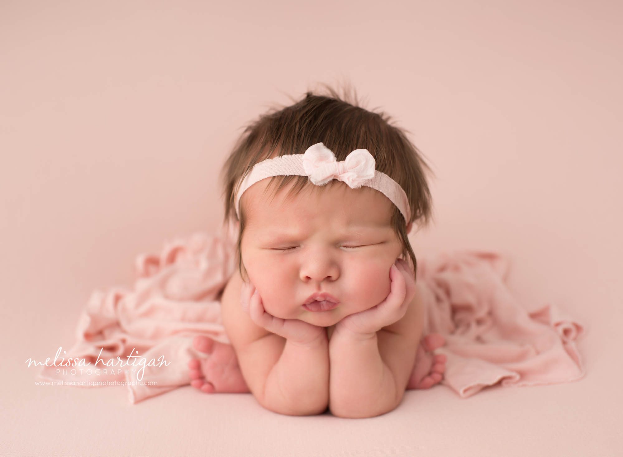 newborn baby girl posed froggy pose wearing bow headband and pink draping wrap puckered lips