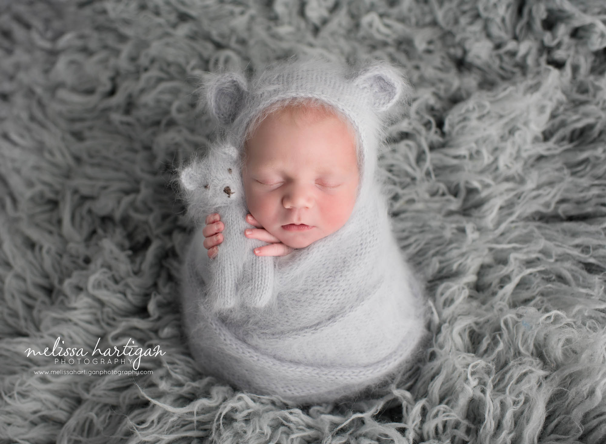 newborn baby boy wrapped in light bluegray knitted wrap wearing matching bear bonnet and holding knitted teddy